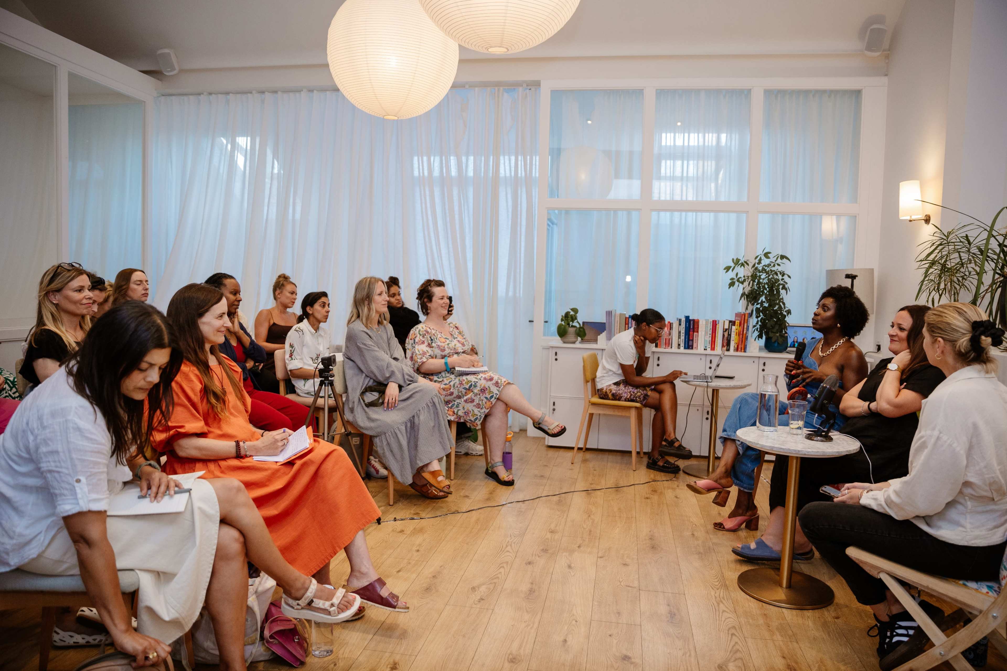 A group of women is seated in a bright, airy room, engaged in discussion, while a speaker addresses them from the front.