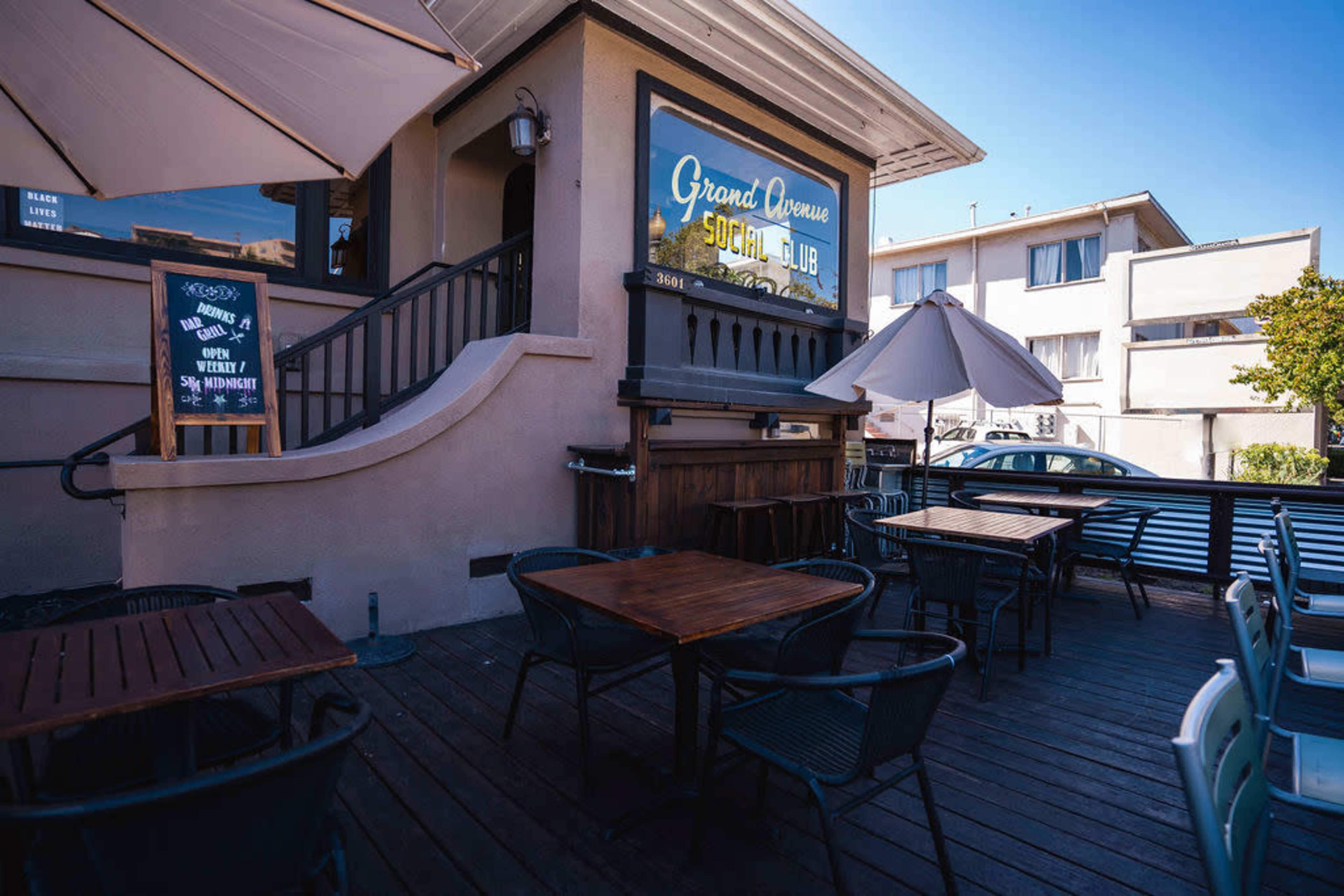 The image shows the exterior of a building with a sign that reads "Grand Avenue Social Club," featuring outdoor seating with tables and umbrellas.