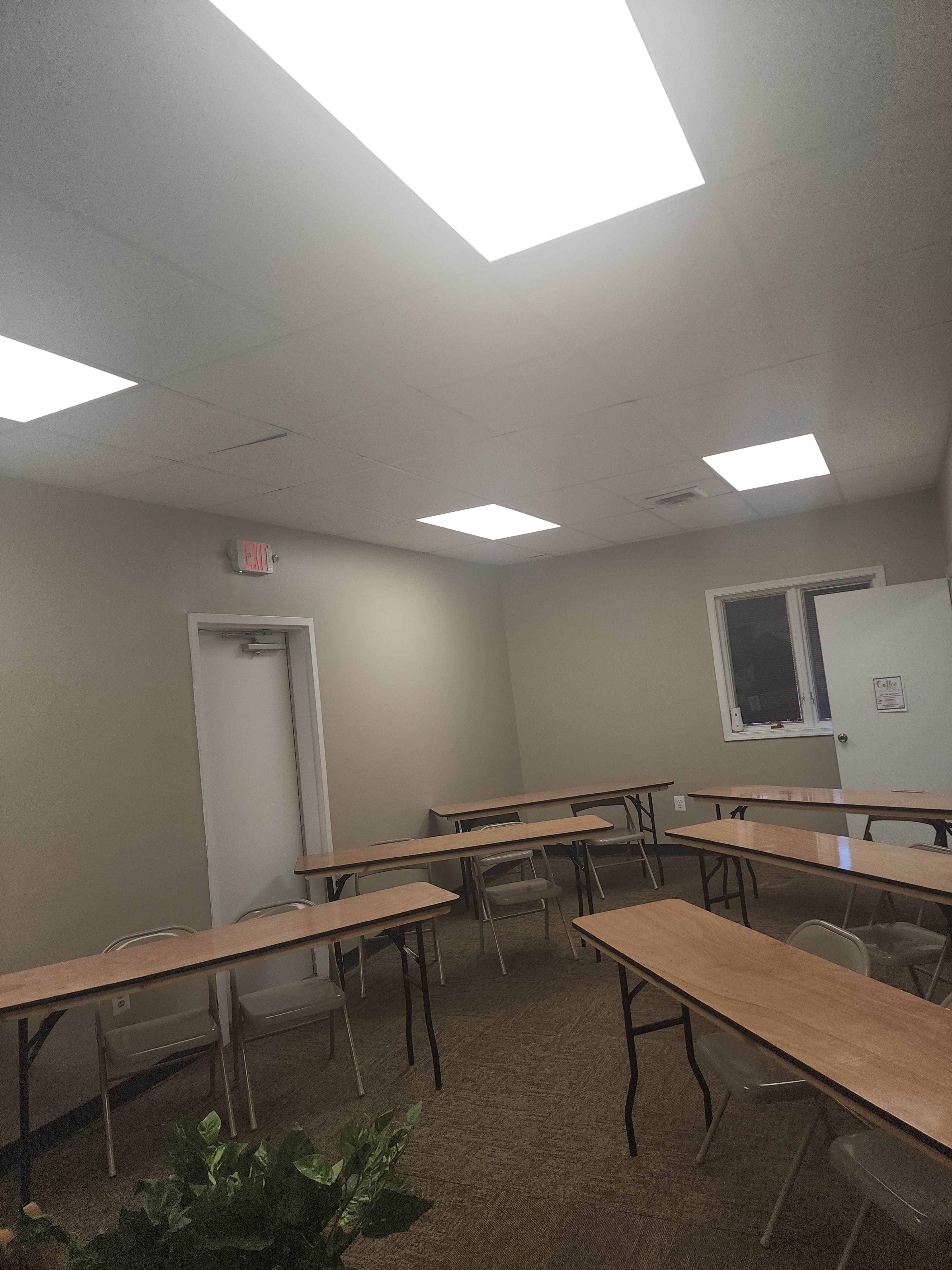 A small meeting room with several tables arranged in a U-shape, beige walls, and bright fluorescent lighting.
