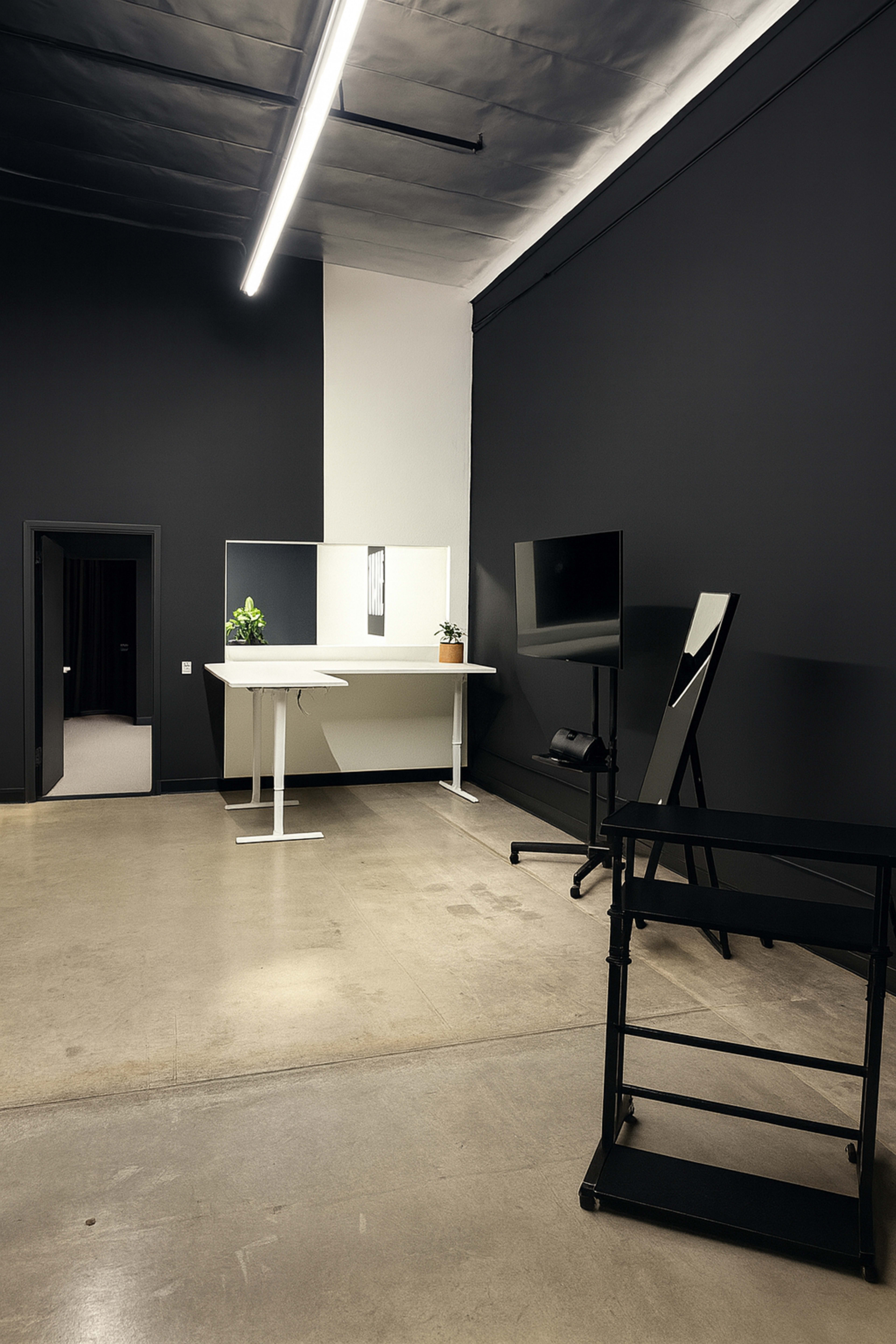 A minimalist room features a black wall, a white table, a mirror, a potted plant, and an empty easel beside a television on a stand.