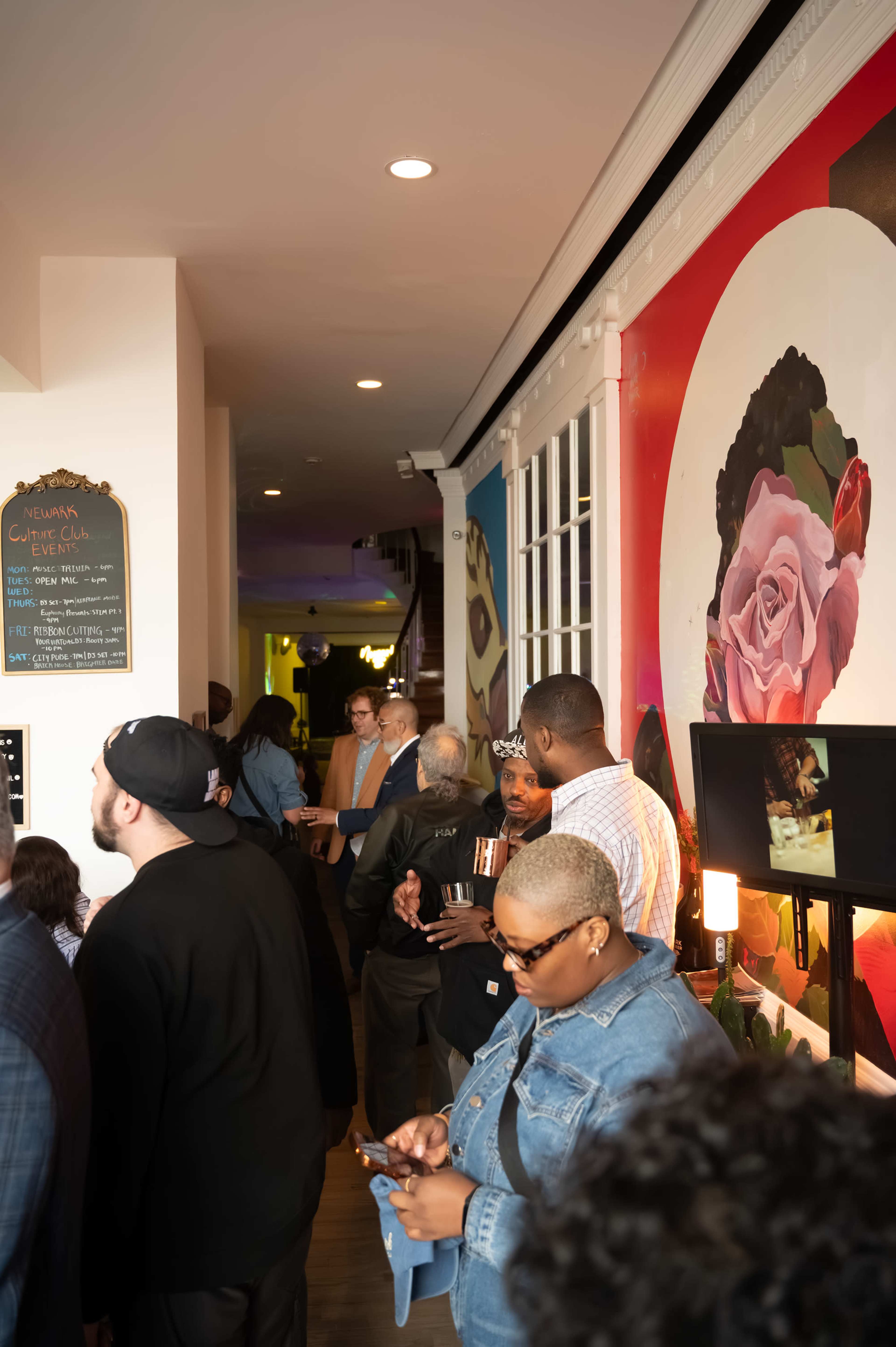 A crowd of people stands in a narrow hallway lined with colorful artwork and a chalkboard menu.