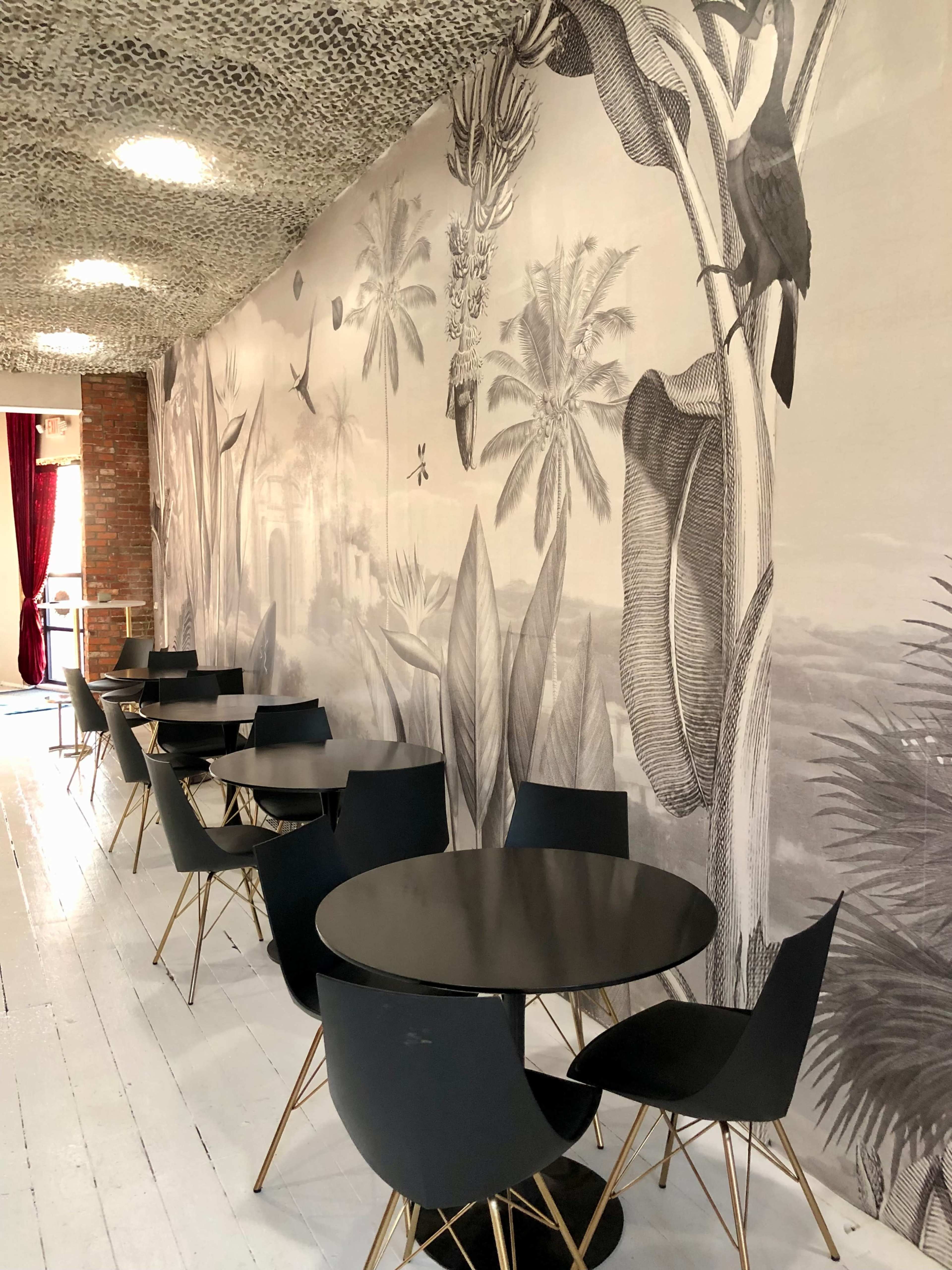 A cafe interior with black tables and chairs arranged on a white wooden floor, featuring a large botanical mural on one wall.