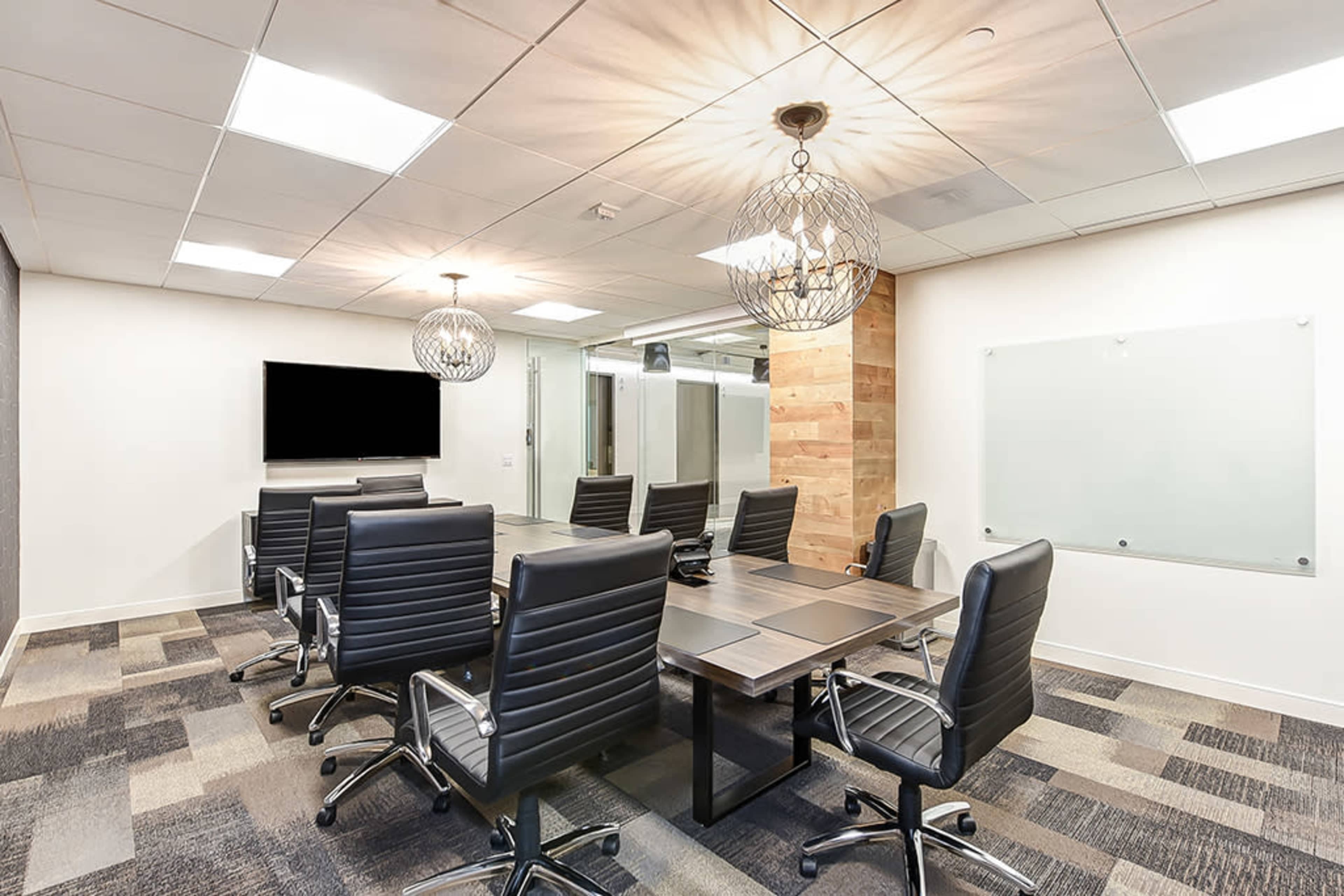A modern conference room features a long wooden table surrounded by black leather chairs, with pendant lighting and a large screen mounted on the wall.