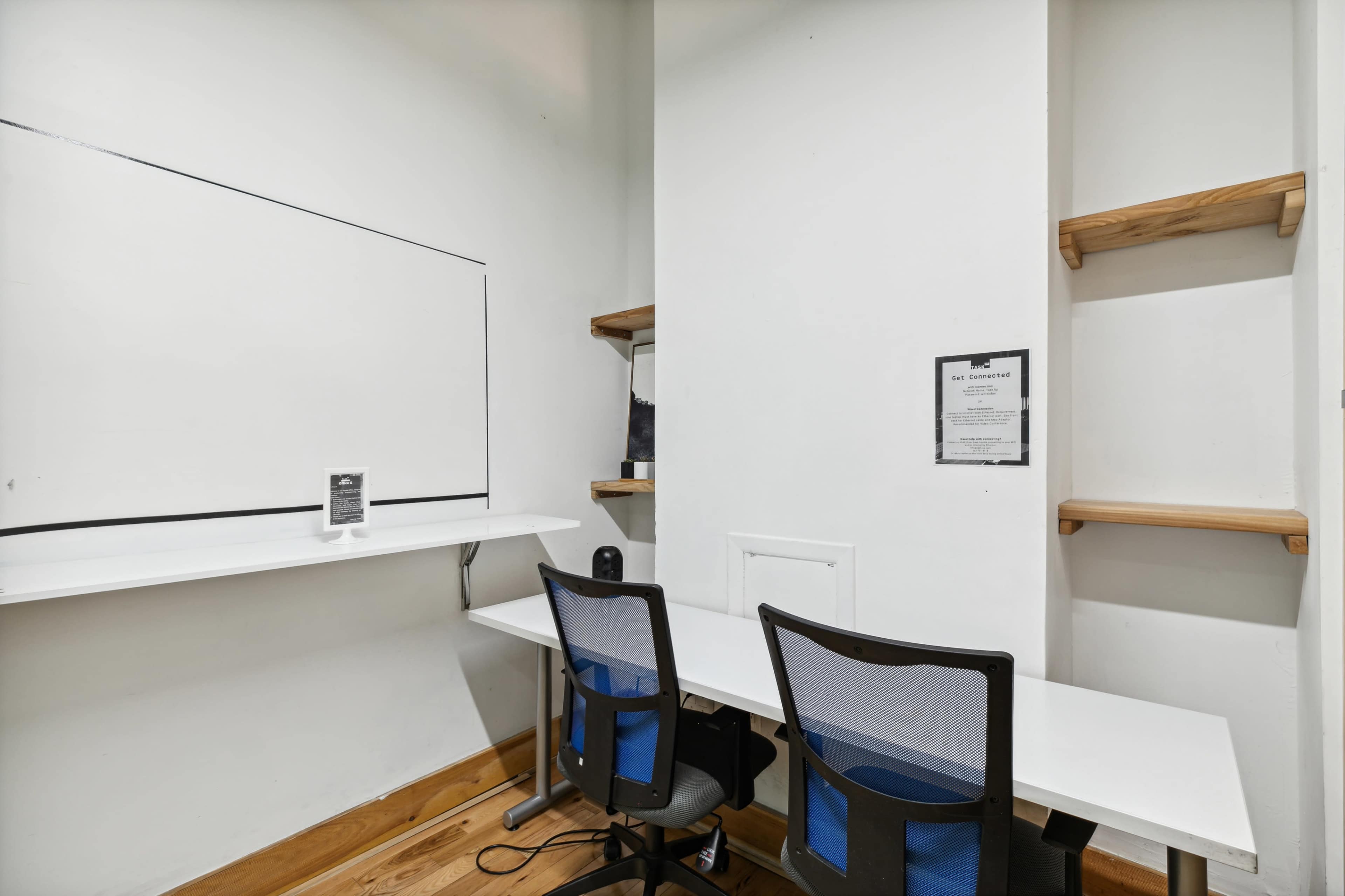A small, minimalistic workspace featuring two black mesh office chairs at a white desk, with a whiteboard and shelves on the wall.