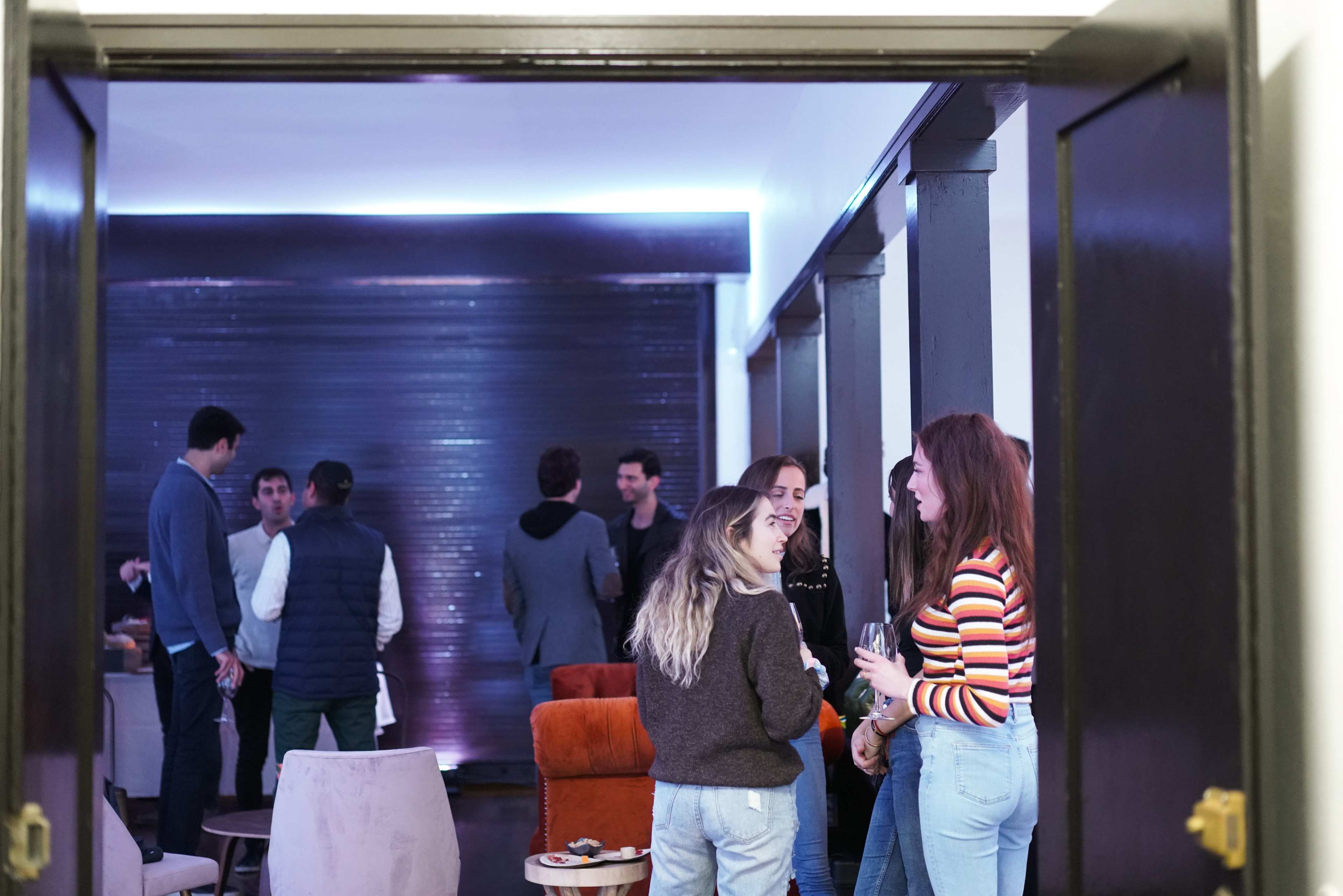 A group of people is socializing in a dimly lit room, with some chatting near the entrance and others standing further back.