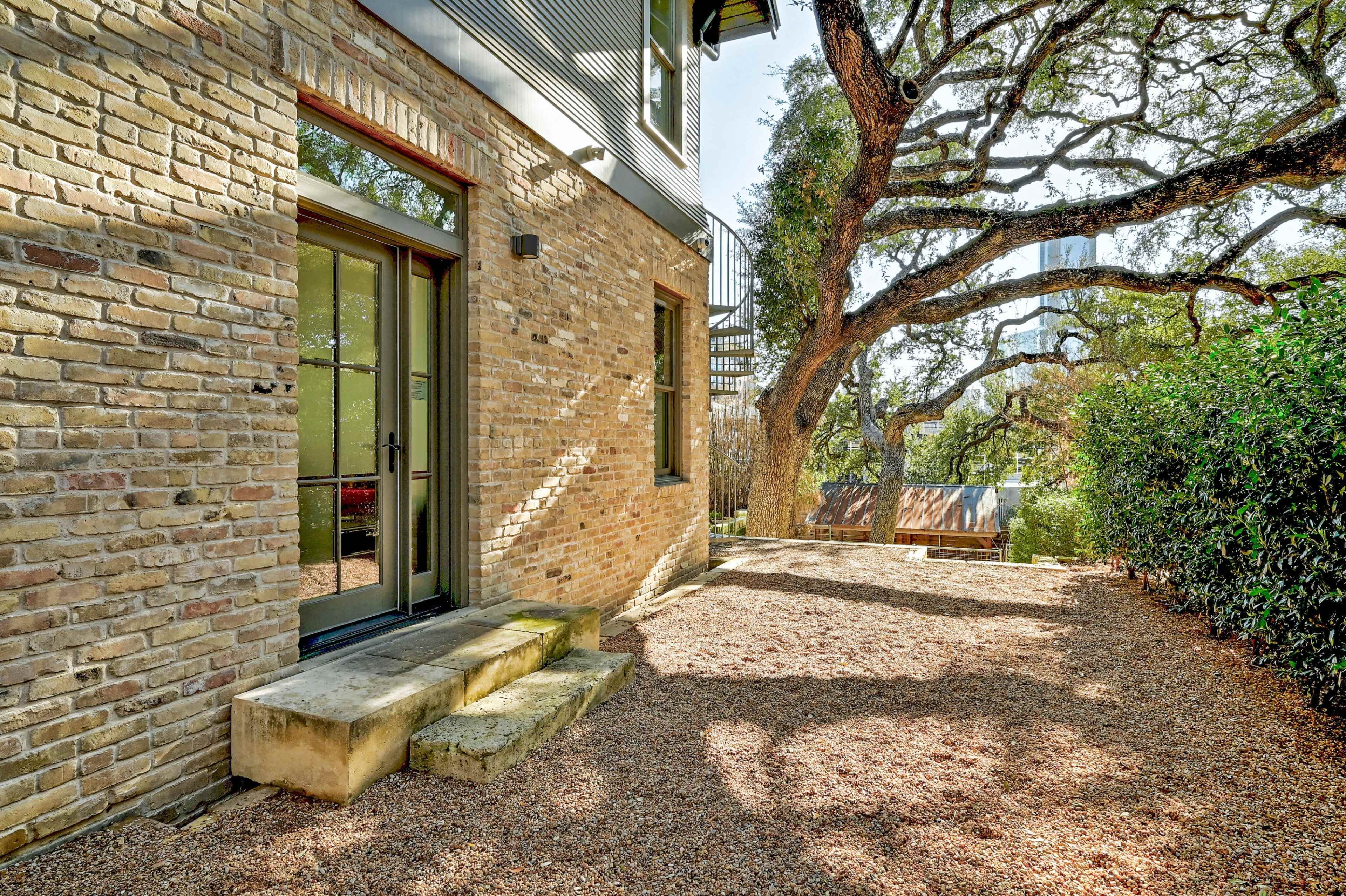 The image shows a brick building with large windows adjacent to a shaded area featuring a gravel path and a large tree.