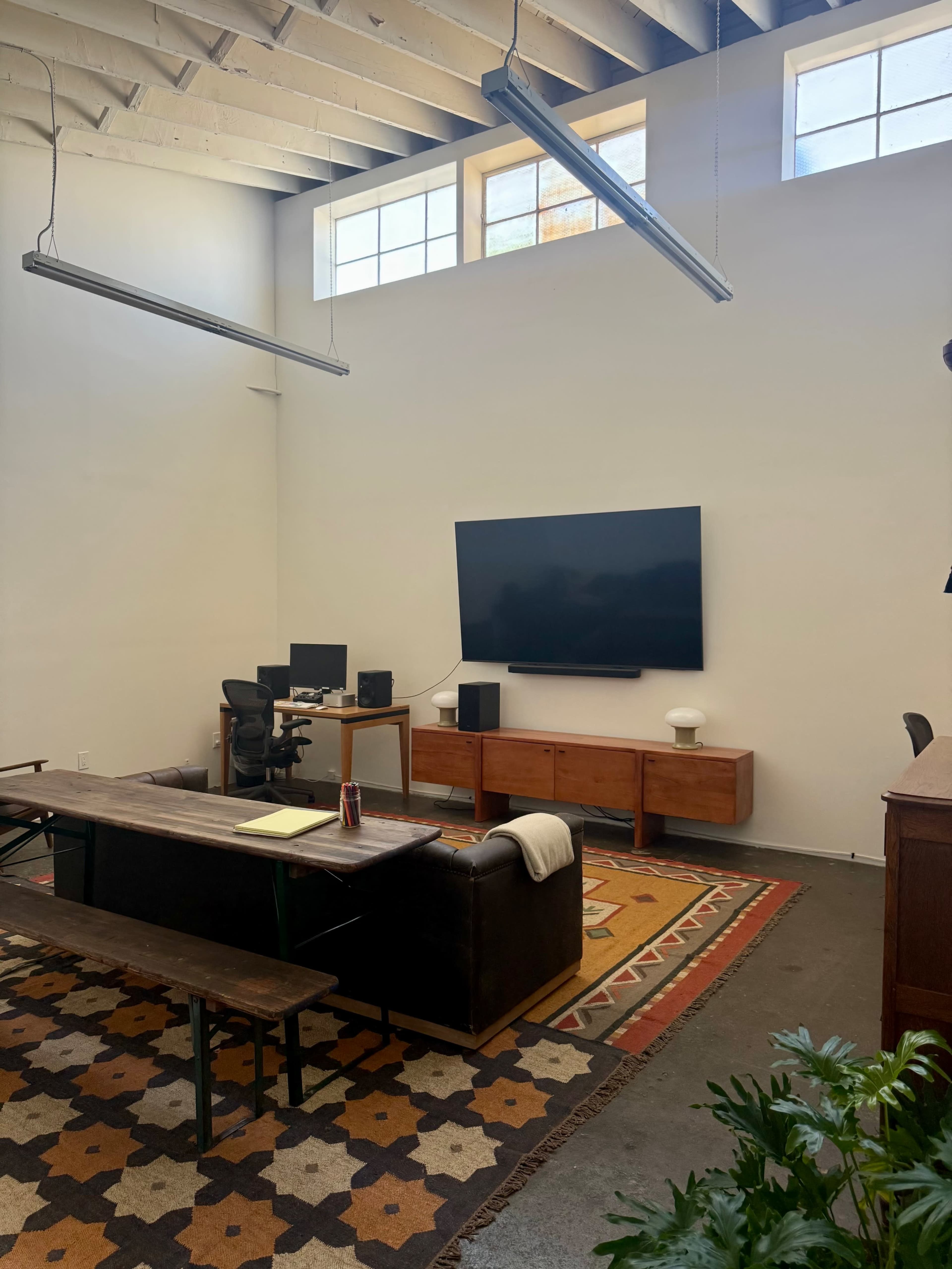 The image shows a spacious, minimalist office with a large wall-mounted television, a wooden table with benches, and a modern desk setup.