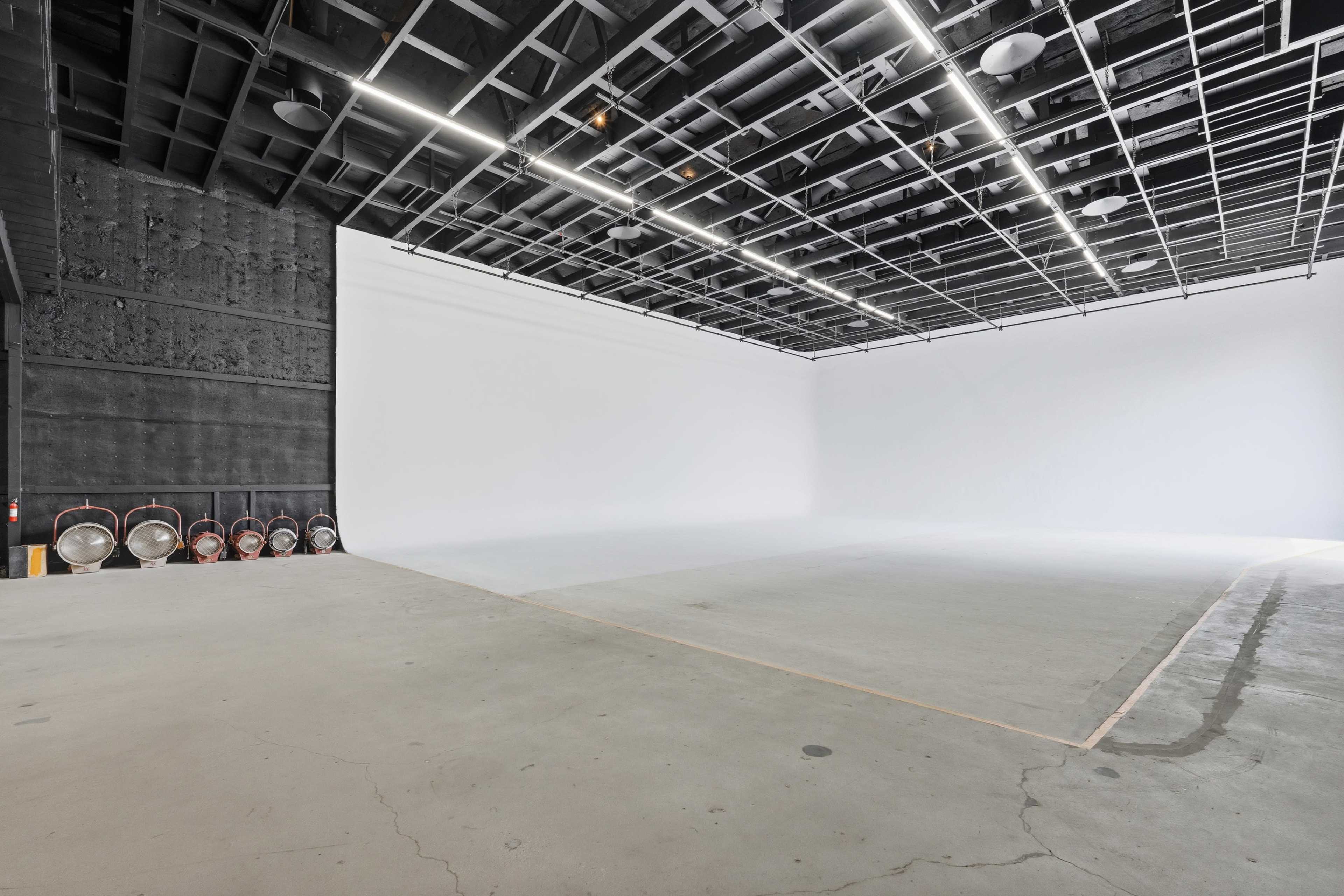 The image shows a large, empty studio space with a white background and a concrete floor, featuring several round lights and a black wall on one side.
