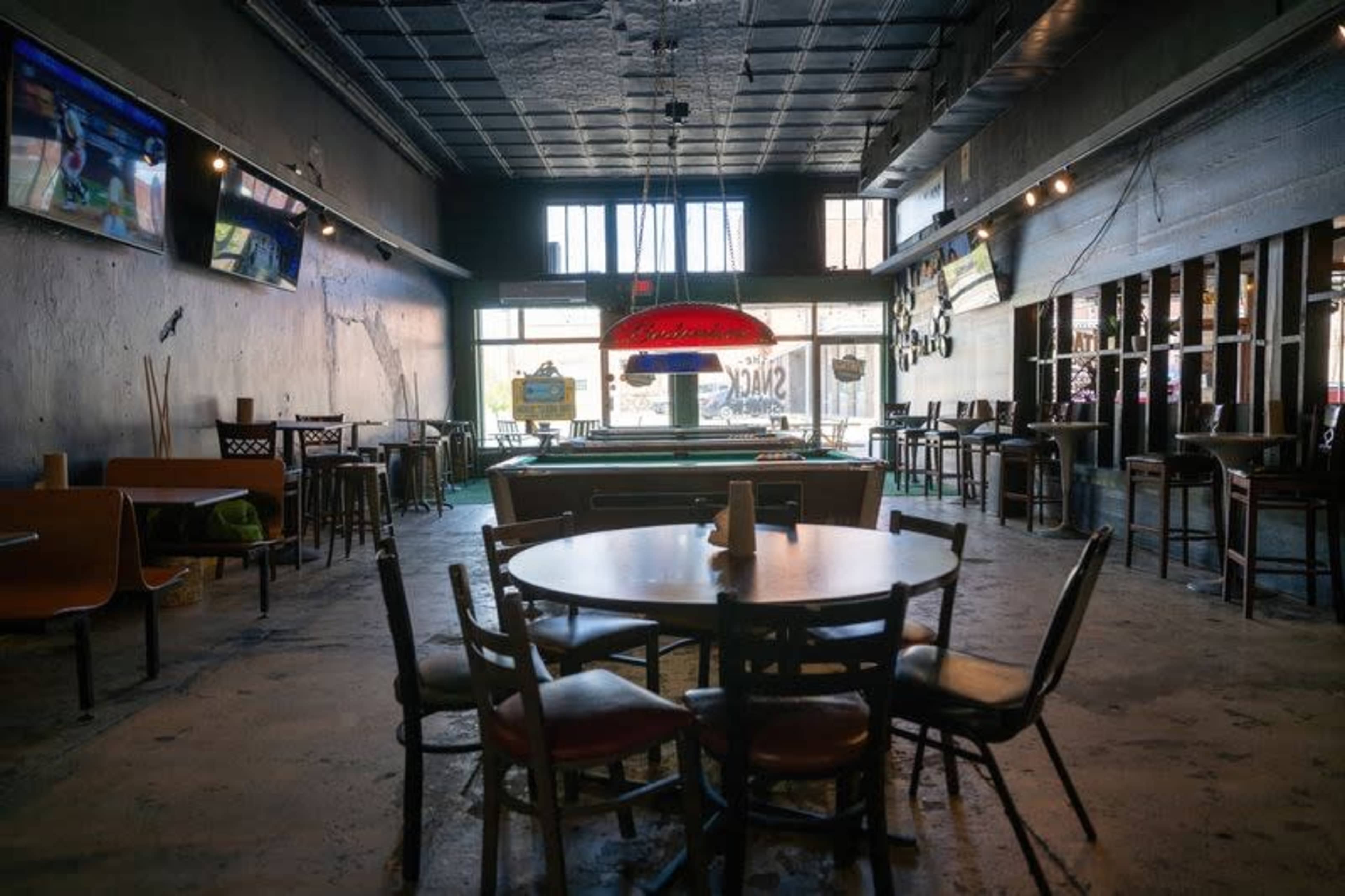 The image shows an empty bar interior with wooden tables and chairs, a pool table, and large windows at the front.