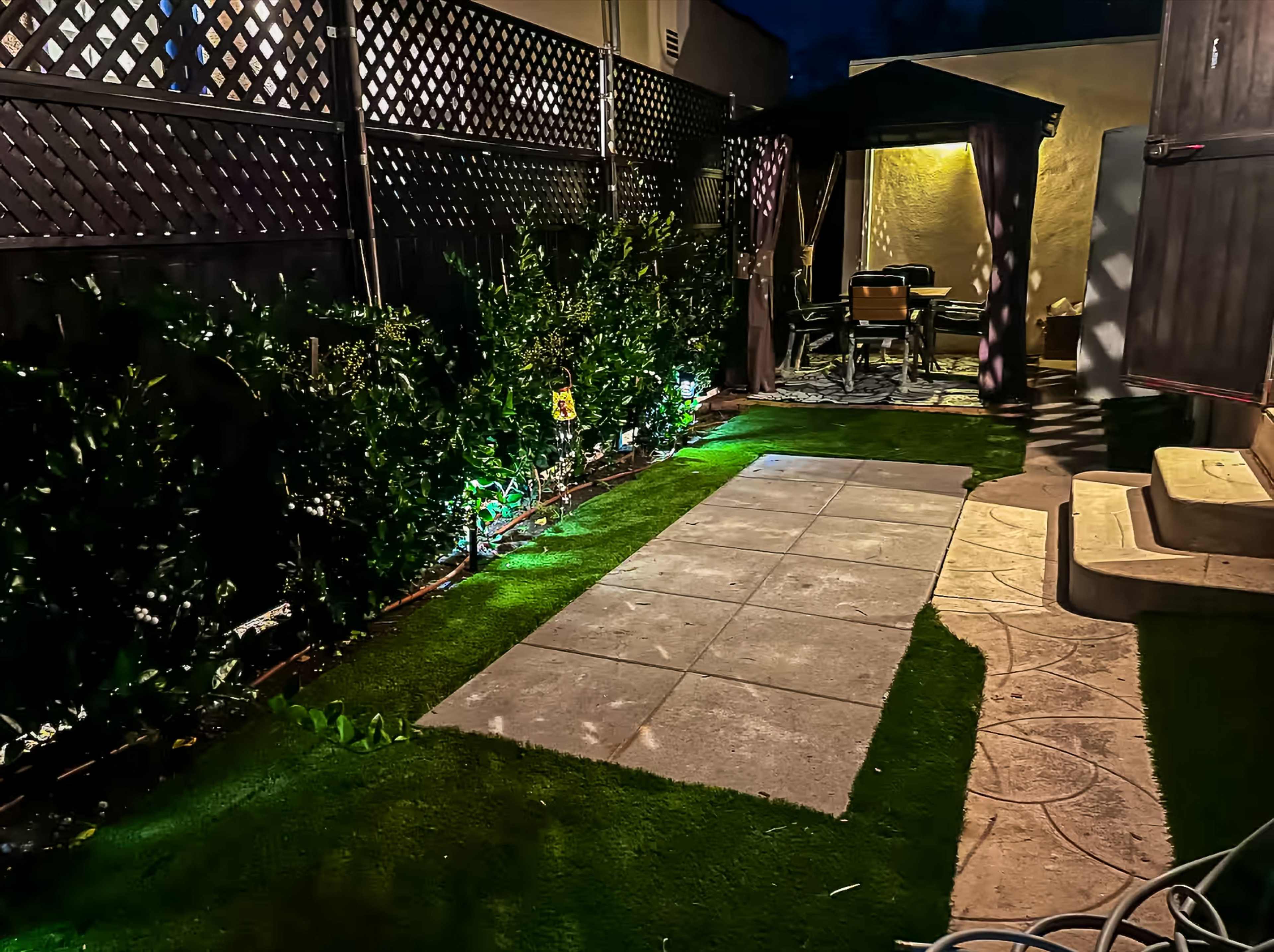 The image depicts a dimly lit backyard featuring a paved walkway, manicured lawn, and a seating area under a canopy.