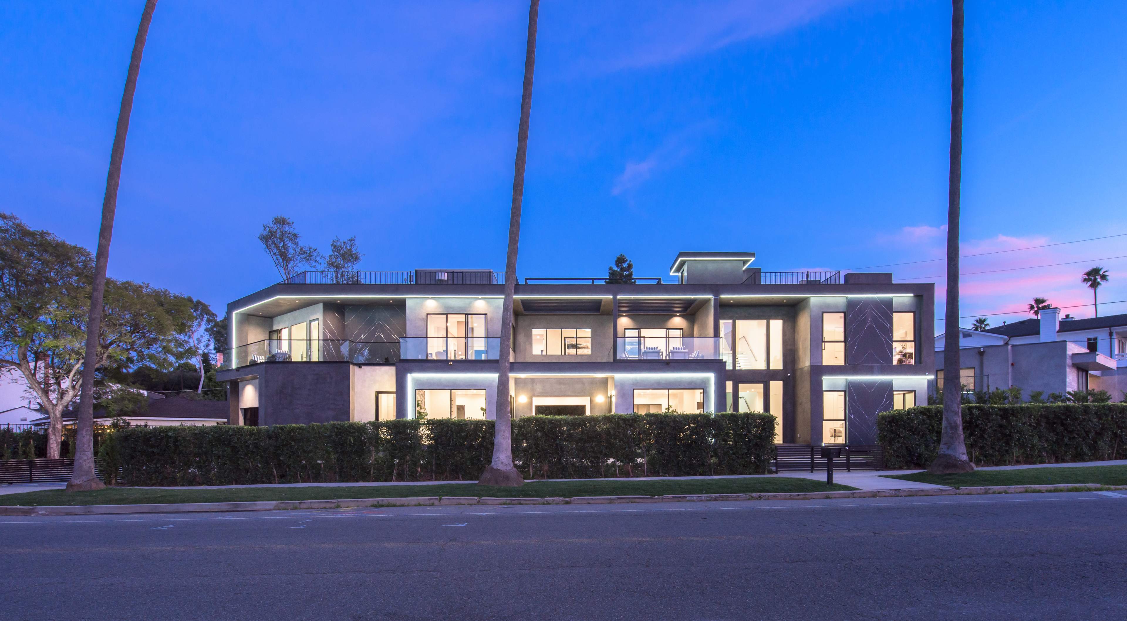 A modern multi-unit building with large windows and a sleek design is situated along a palm tree-lined street at dusk.