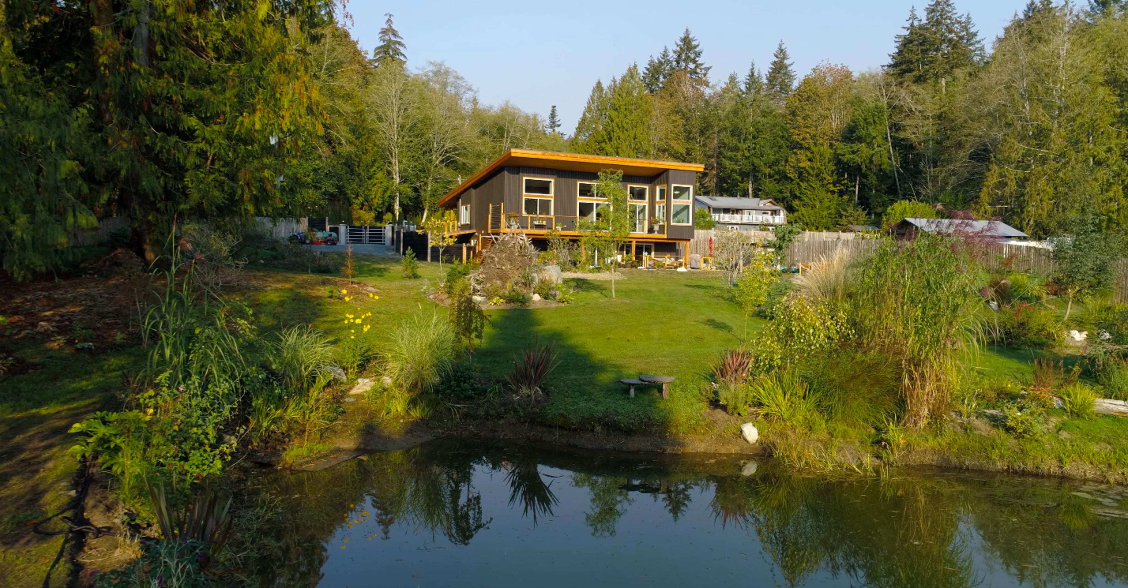 A modern house with large windows is situated on a grassy lawn beside a serene pond, surrounded by trees and landscaping.