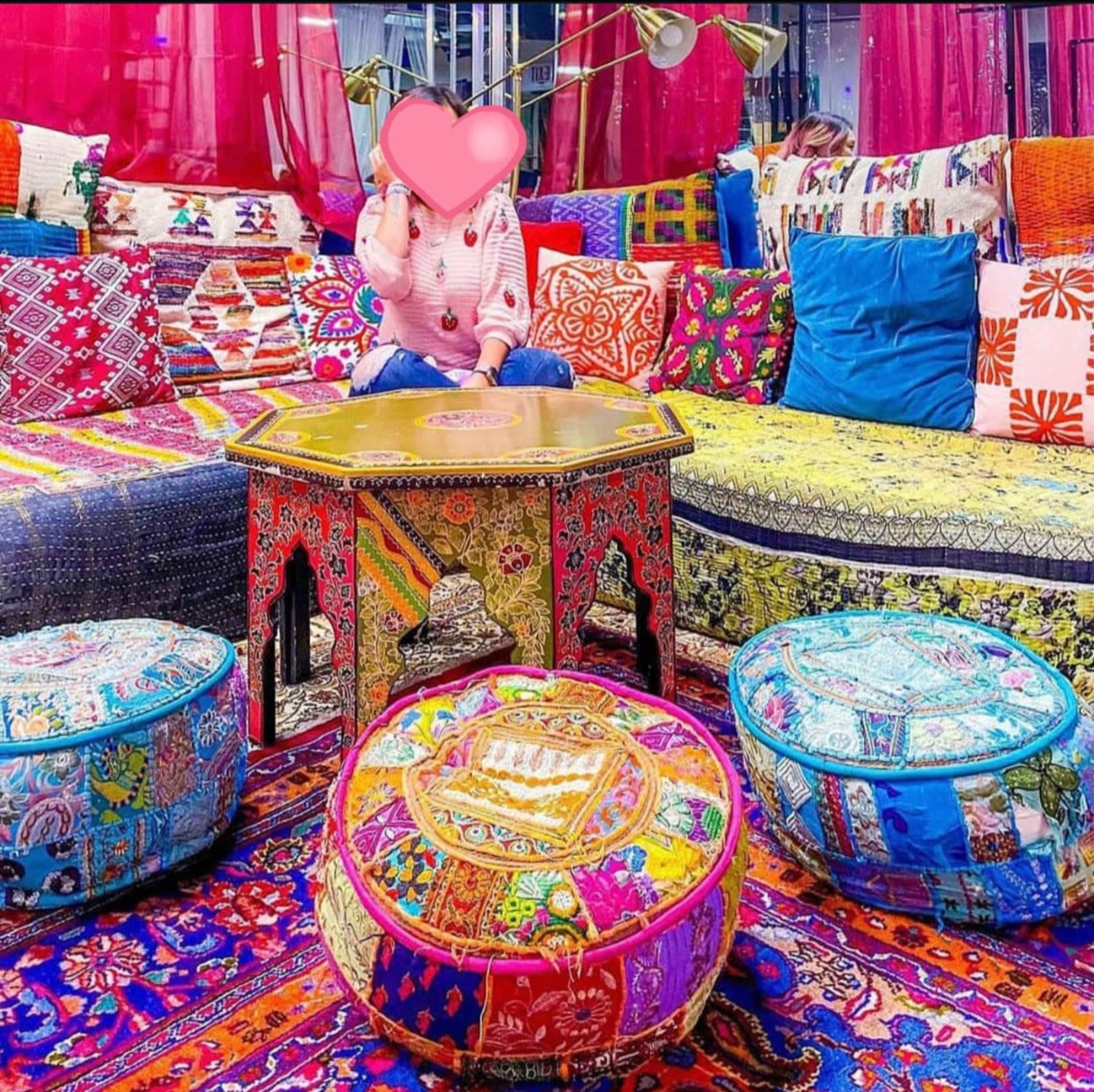 The scene features a vibrant, colorful seating area adorned with patterned cushions, embroidered poufs, and a decorative table.