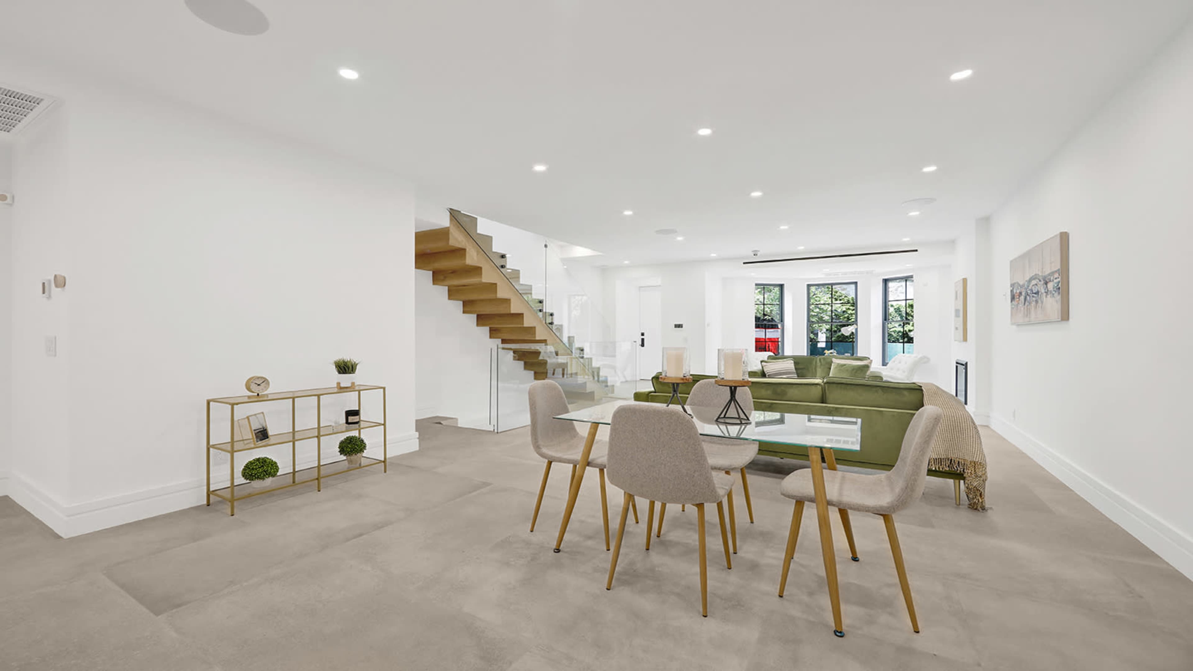 A modern, airy living space features a dining table with chairs, a staircase leading to the upper floor, and a green sofa arranged near large windows.