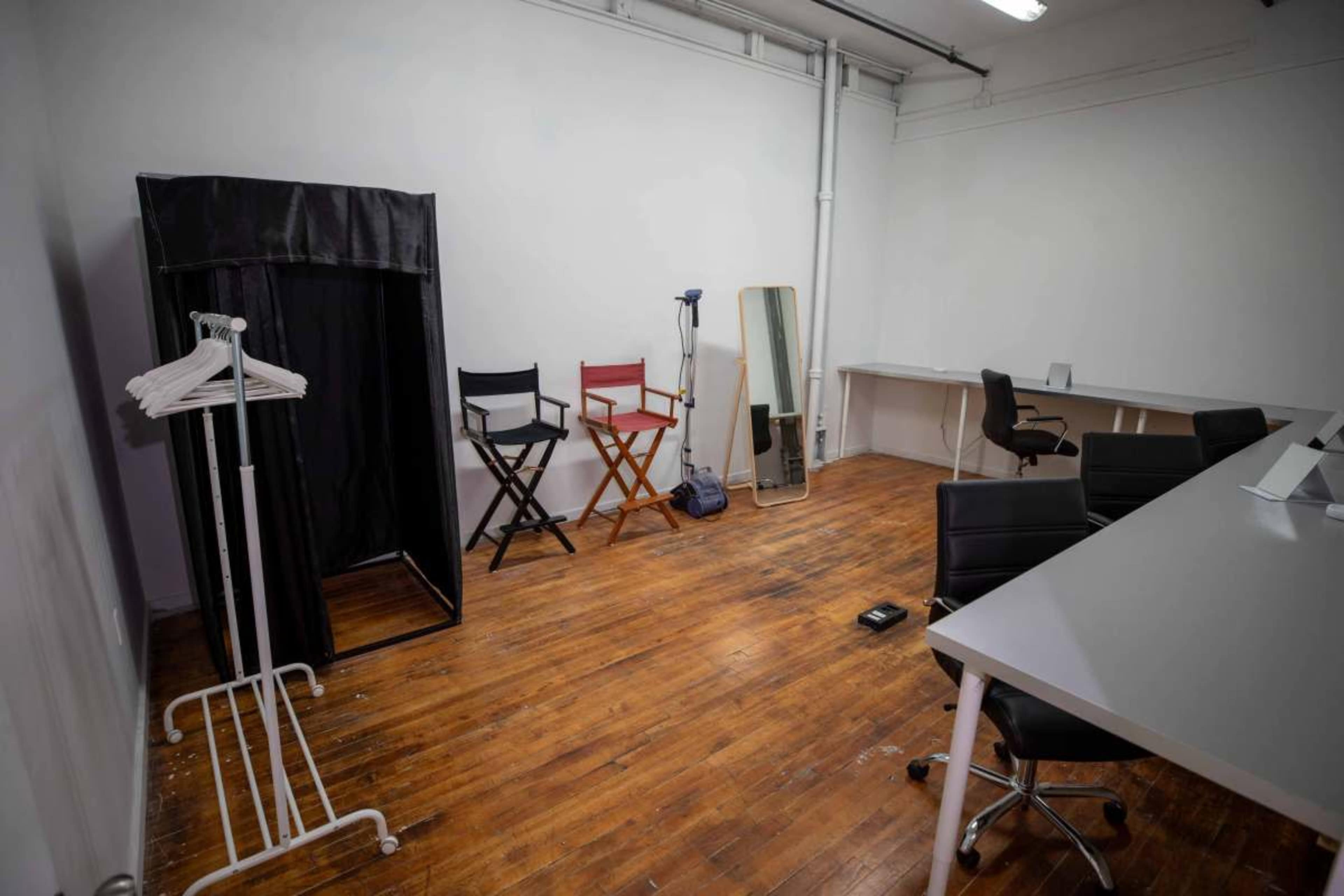 The image shows a sparse, empty room with wooden floors, a portable changing area, two director's chairs, a large mirror, and a desk with chairs.