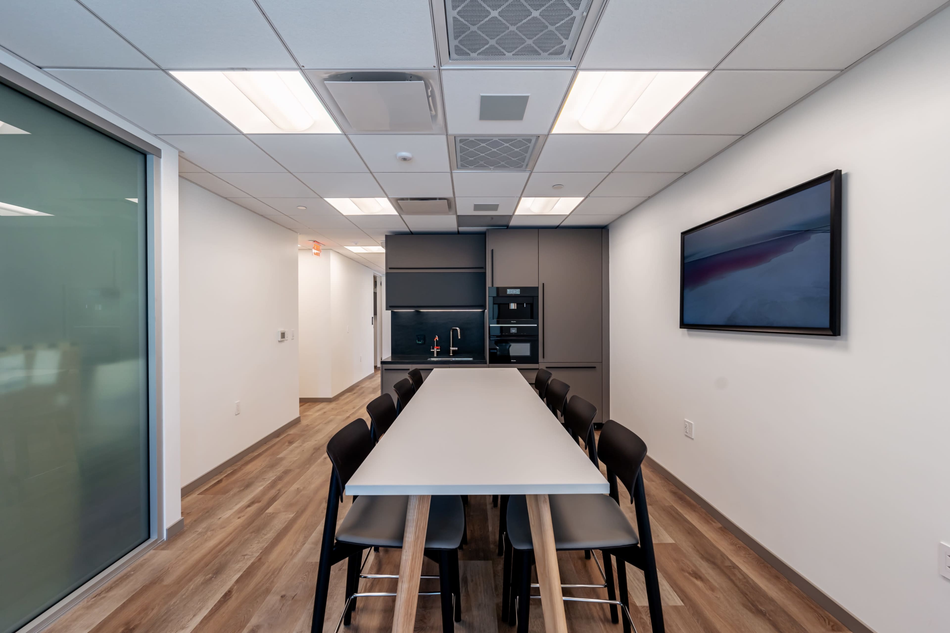 The image shows a modern conference room with a long table surrounded by black chairs, a kitchenette in the back, and a wall-mounted screen.