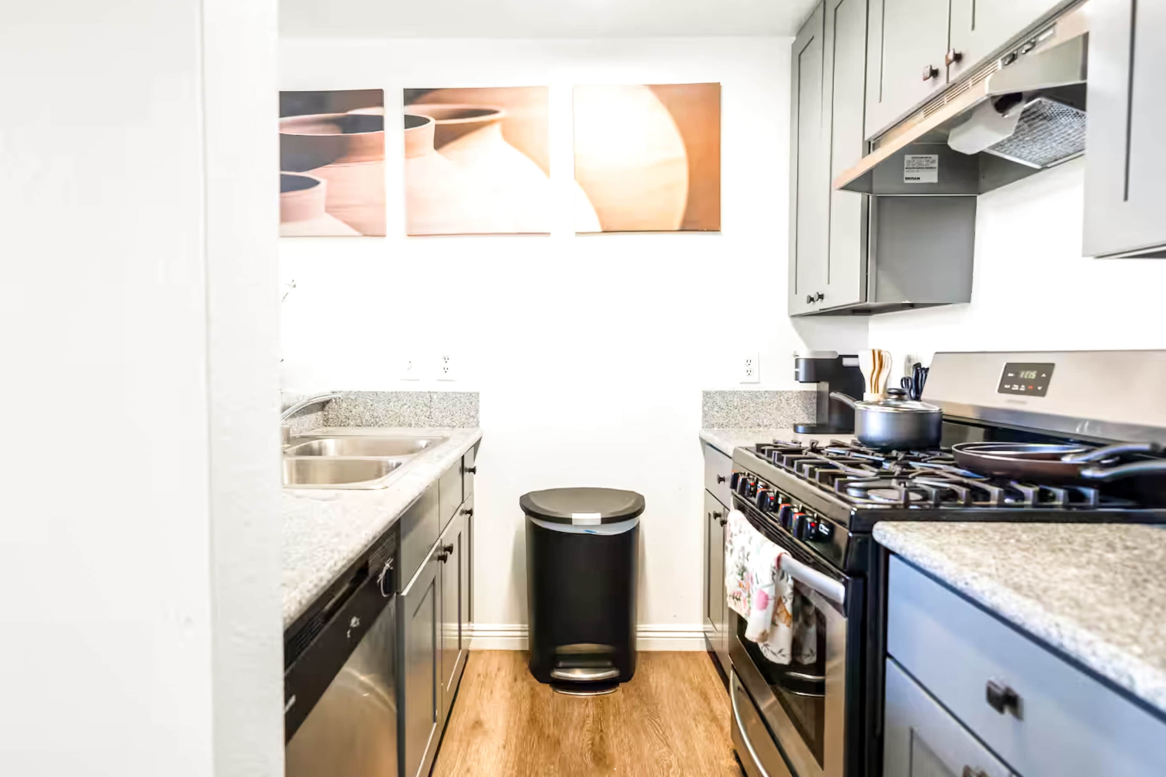 The image shows a modern kitchen featuring gray cabinets, a stainless steel stove, a sink, and a black trash can, with artwork of pottery on the wall.