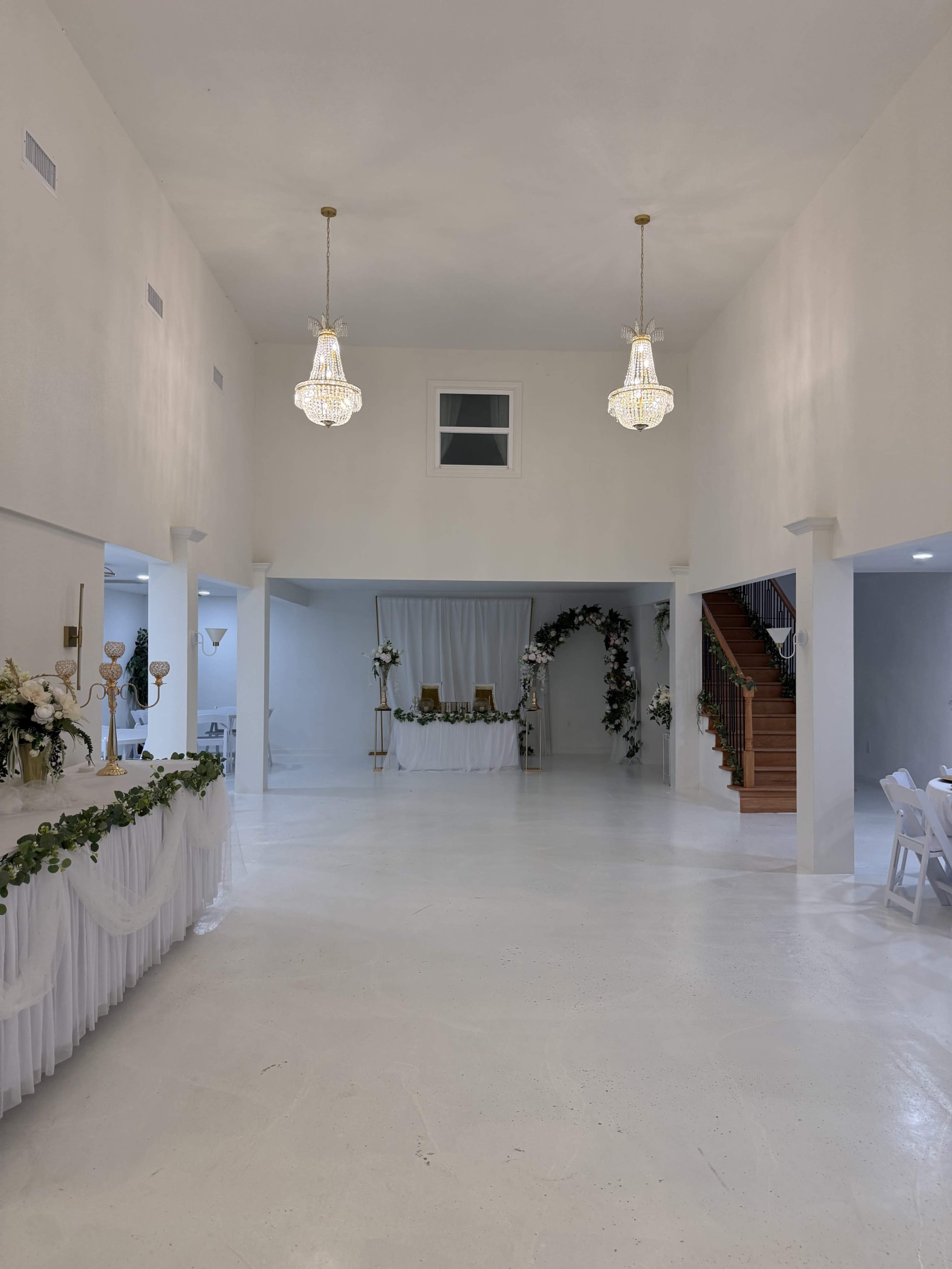 The image shows a spacious, well-lit event hall with chandeliers, a decorated backdrop, and a staircase in the background.