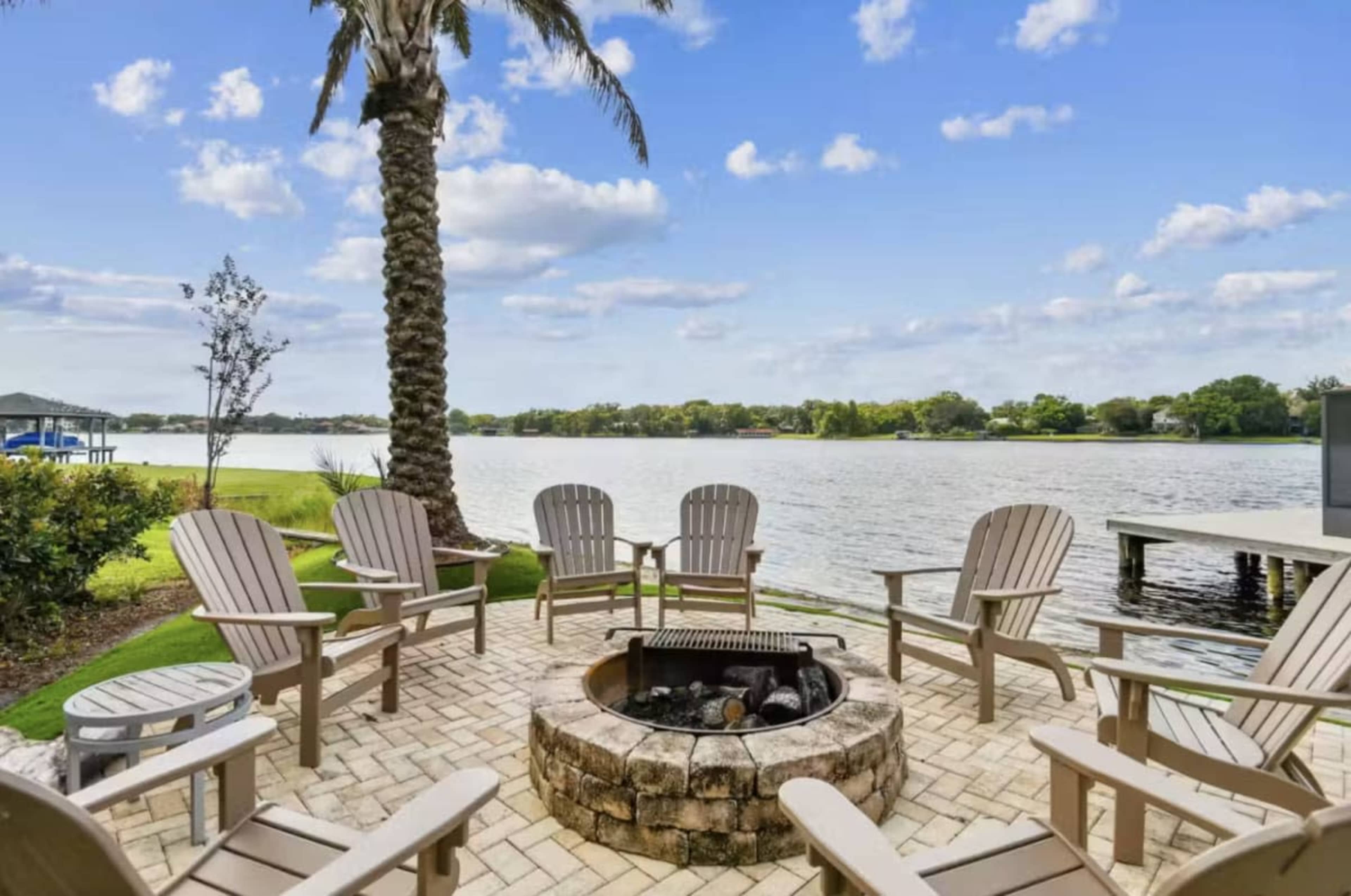 A fire pit surrounded by several wooden chairs is situated next to a lake, with palm trees and green grass in the background.
