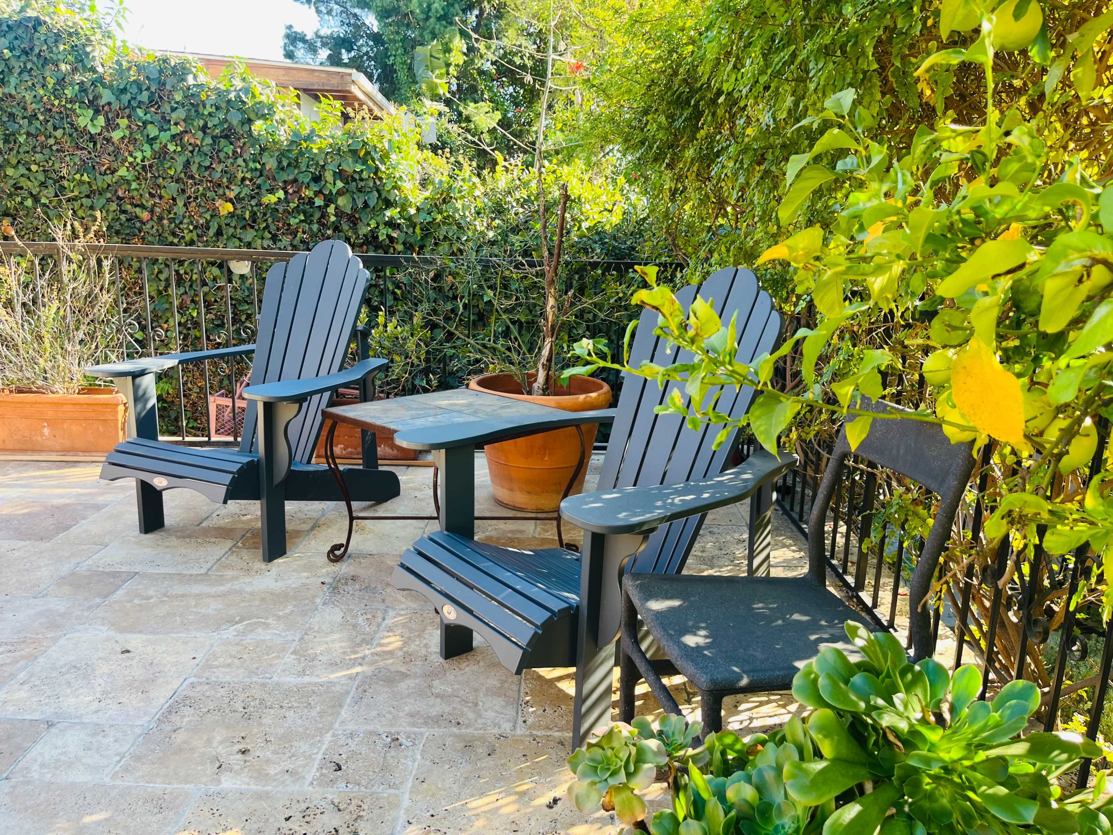 The image shows a outdoor seating area with two adirondack chairs and a small table surrounded by green plants and a garden.