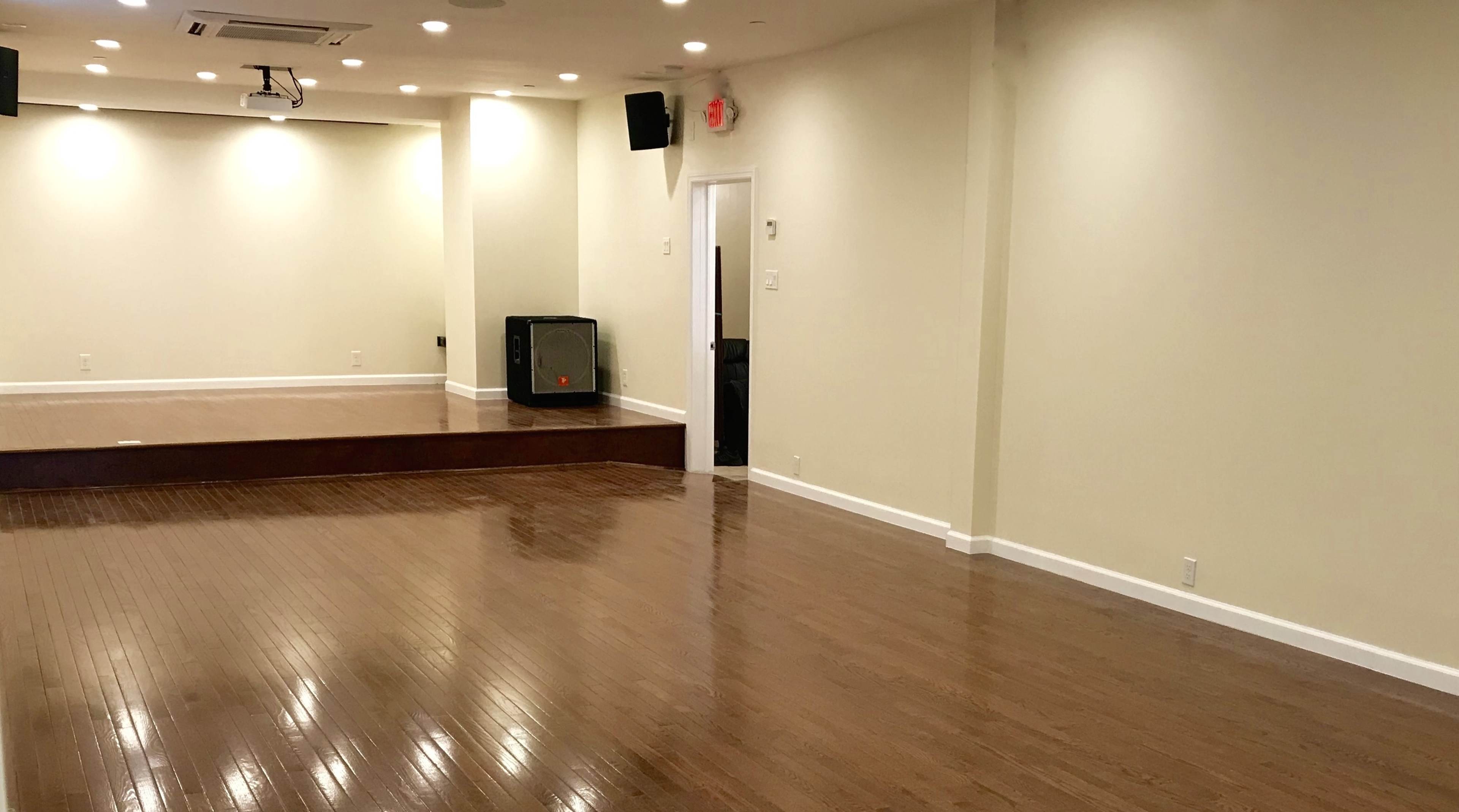 A large, empty room with polished wooden floors, a slight stage in the front, and minimal lighting.