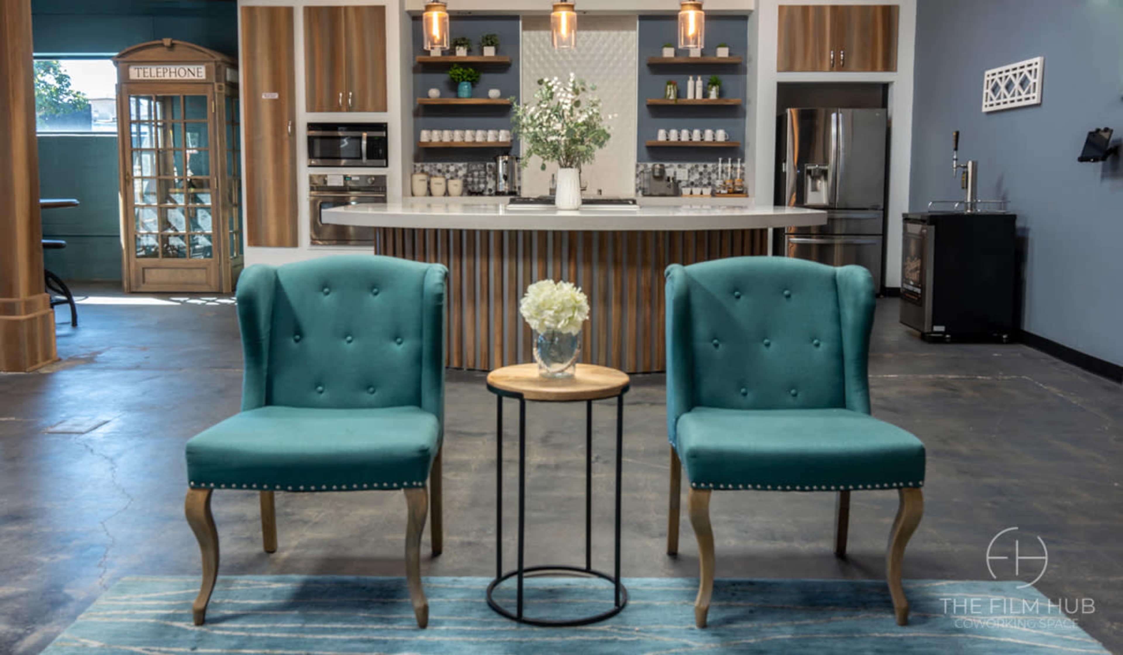 Two teal chairs flanking a small side table with a flower vase, set in a modern kitchen space with a counter and appliances in the background.