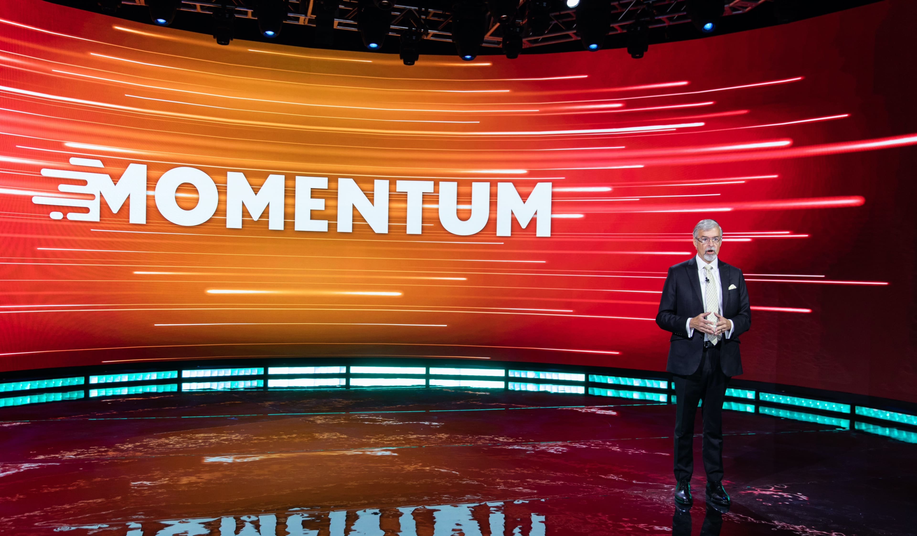 A presenter stands on a stage in front of a large screen displaying the word "MOMENTUM" with dynamic, colorful graphics.