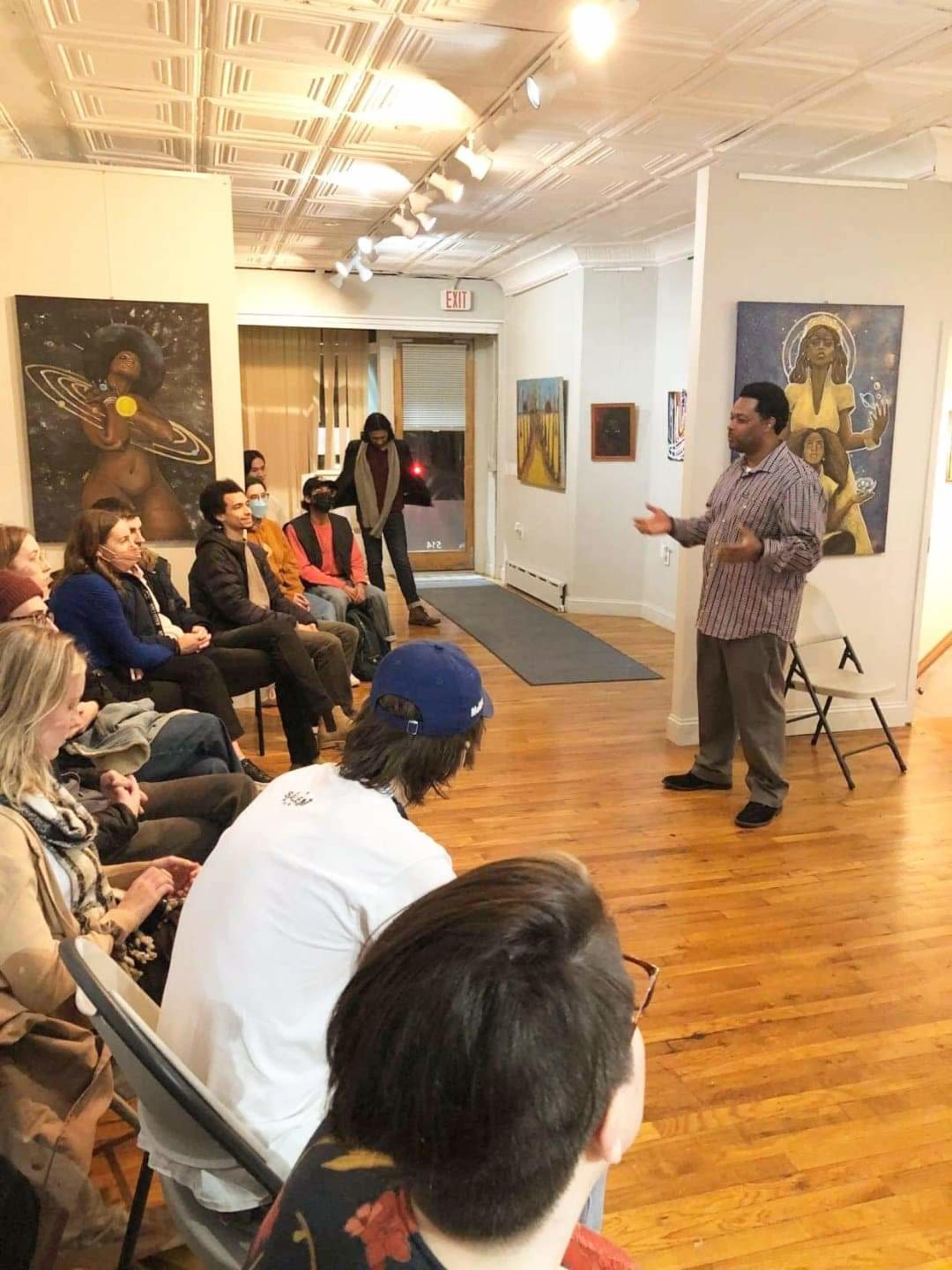 A speaker addresses an audience seated in a gallery filled with artwork on the walls.