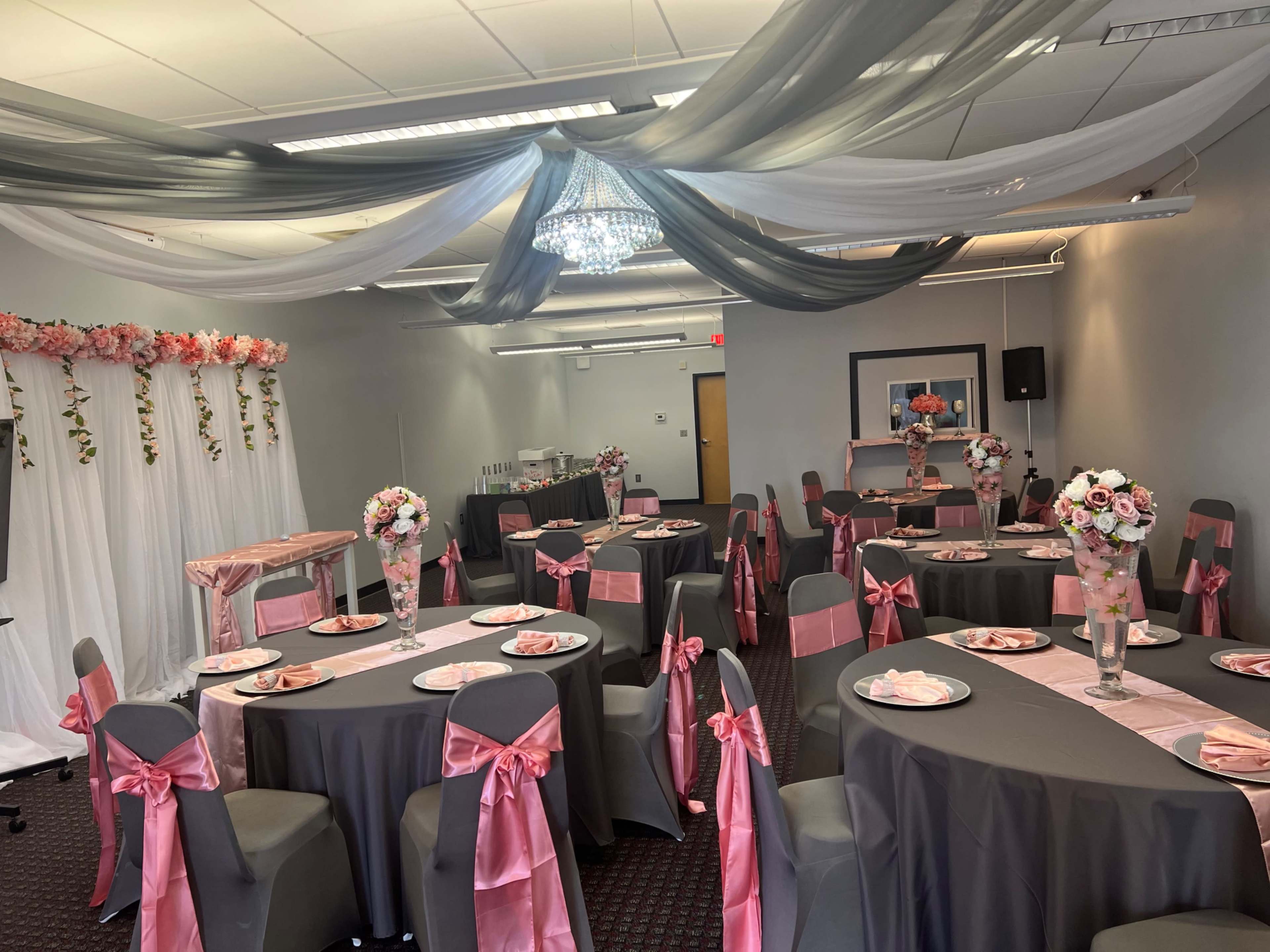 A decorated event space with tables set for a formal gathering, featuring gray tablecloths and pink sashes, floral centerpieces, and draped fabric overhead.