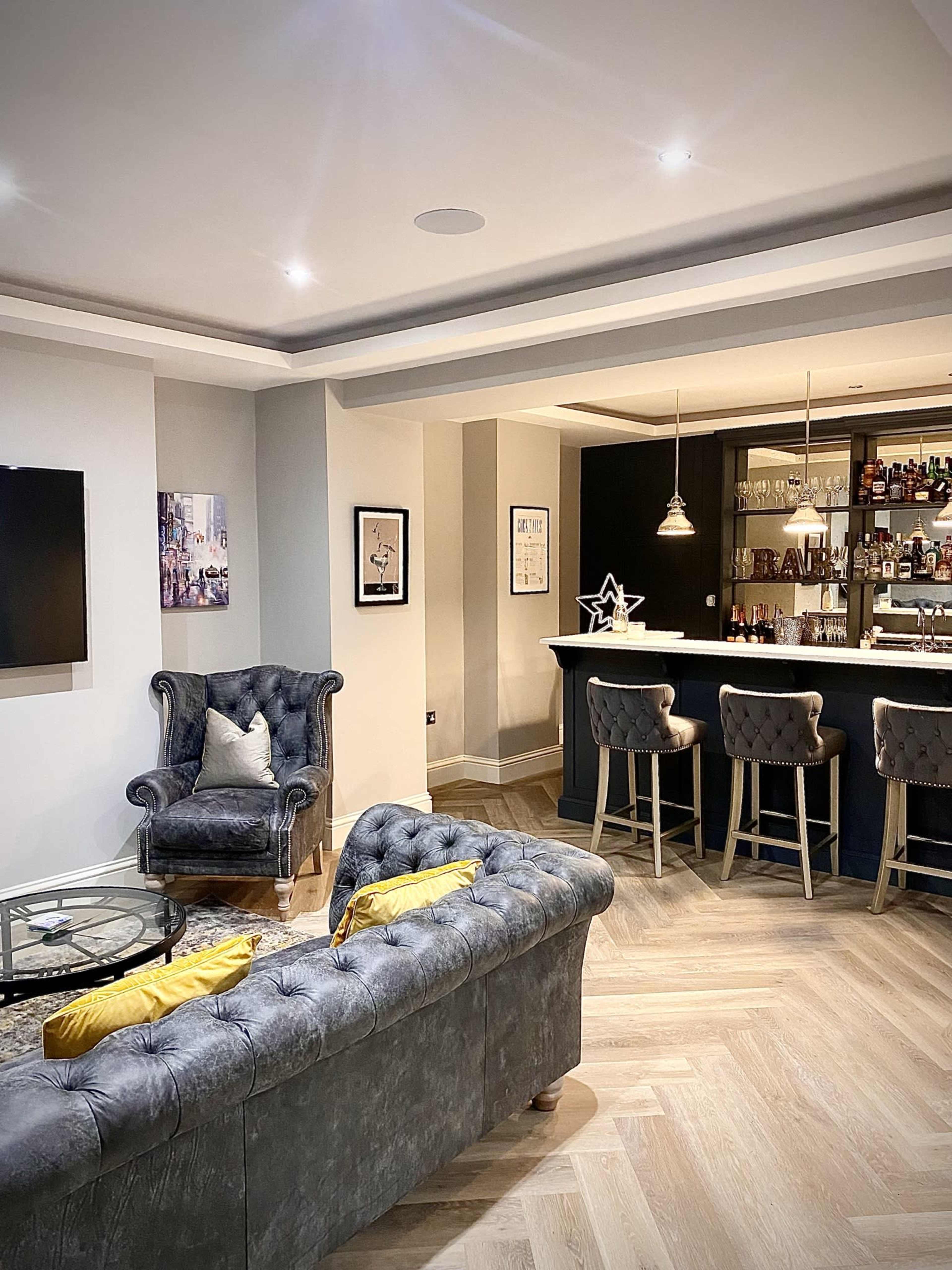 The image shows a modern lounge area featuring a gray tufted sofa, a glass coffee table, and a home bar with high stools and a well-stocked shelf.