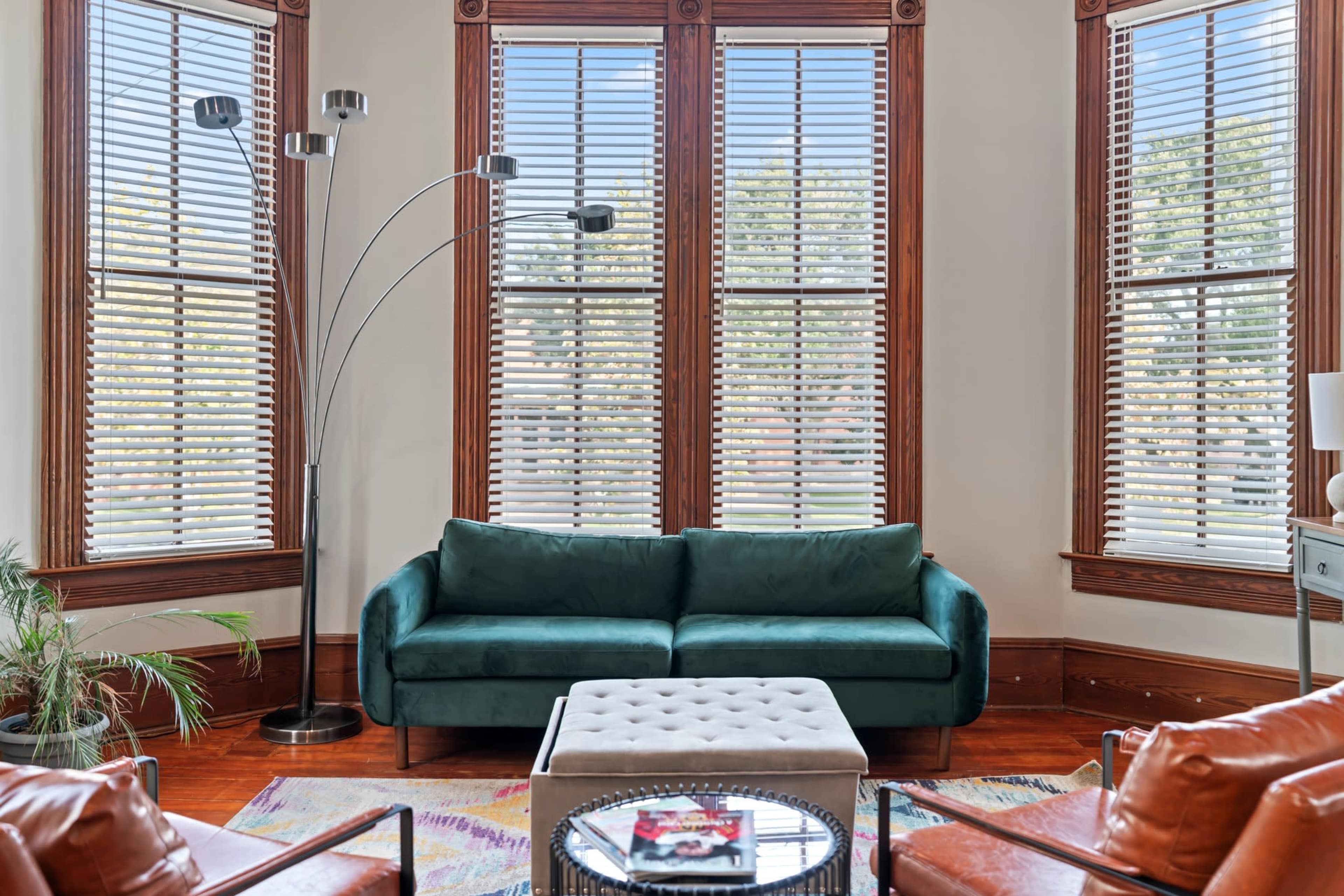 A green sofa sits in a bright room with large windows and wooden blinds, accompanied by a light-colored ottoman and two brown armchairs.