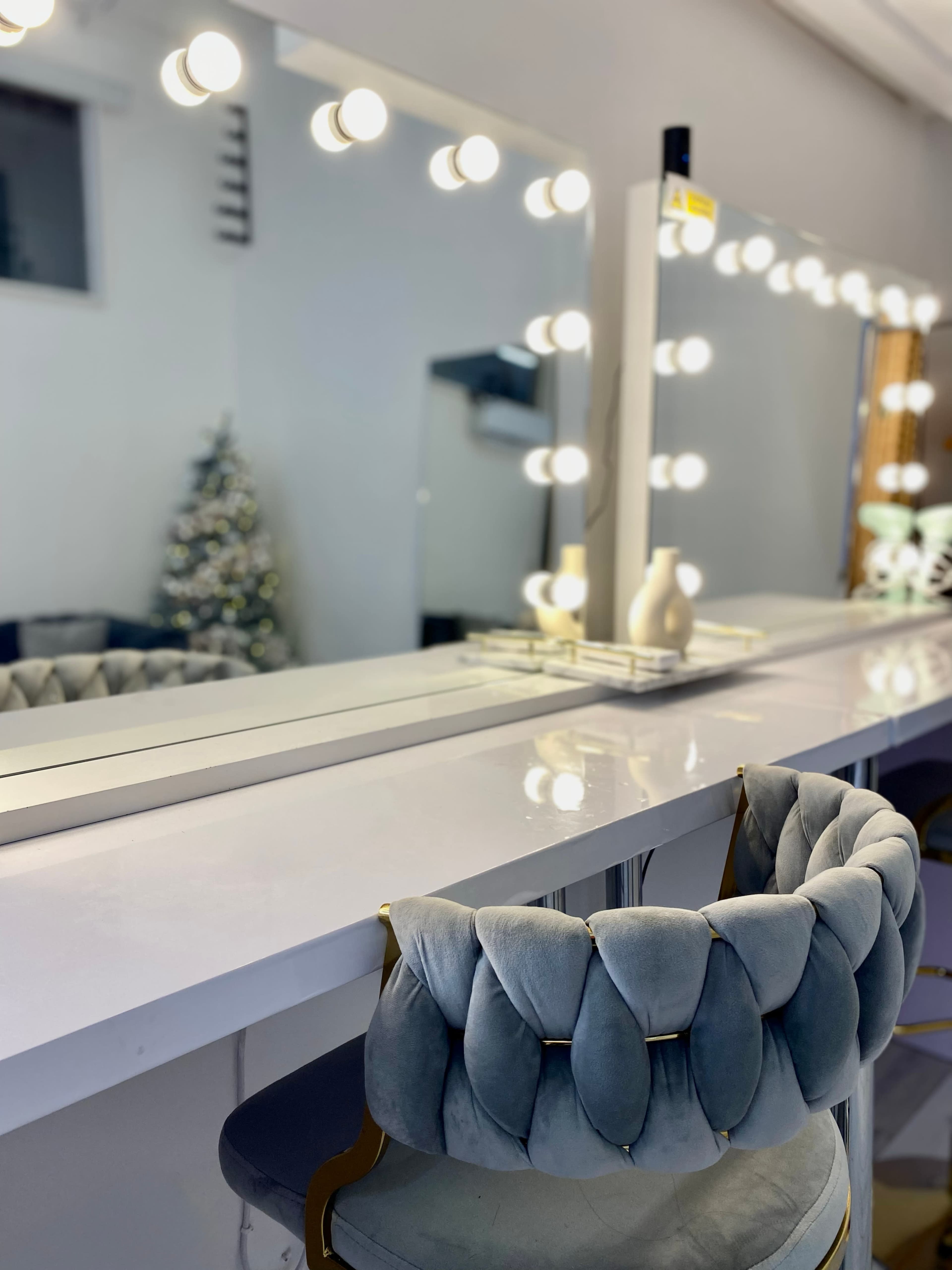 A vanity area features a long mirror with bright circular lights and a plush, tufted chair beside it, with a decorative vase on the countertop and a Christmas tree visible in the background.