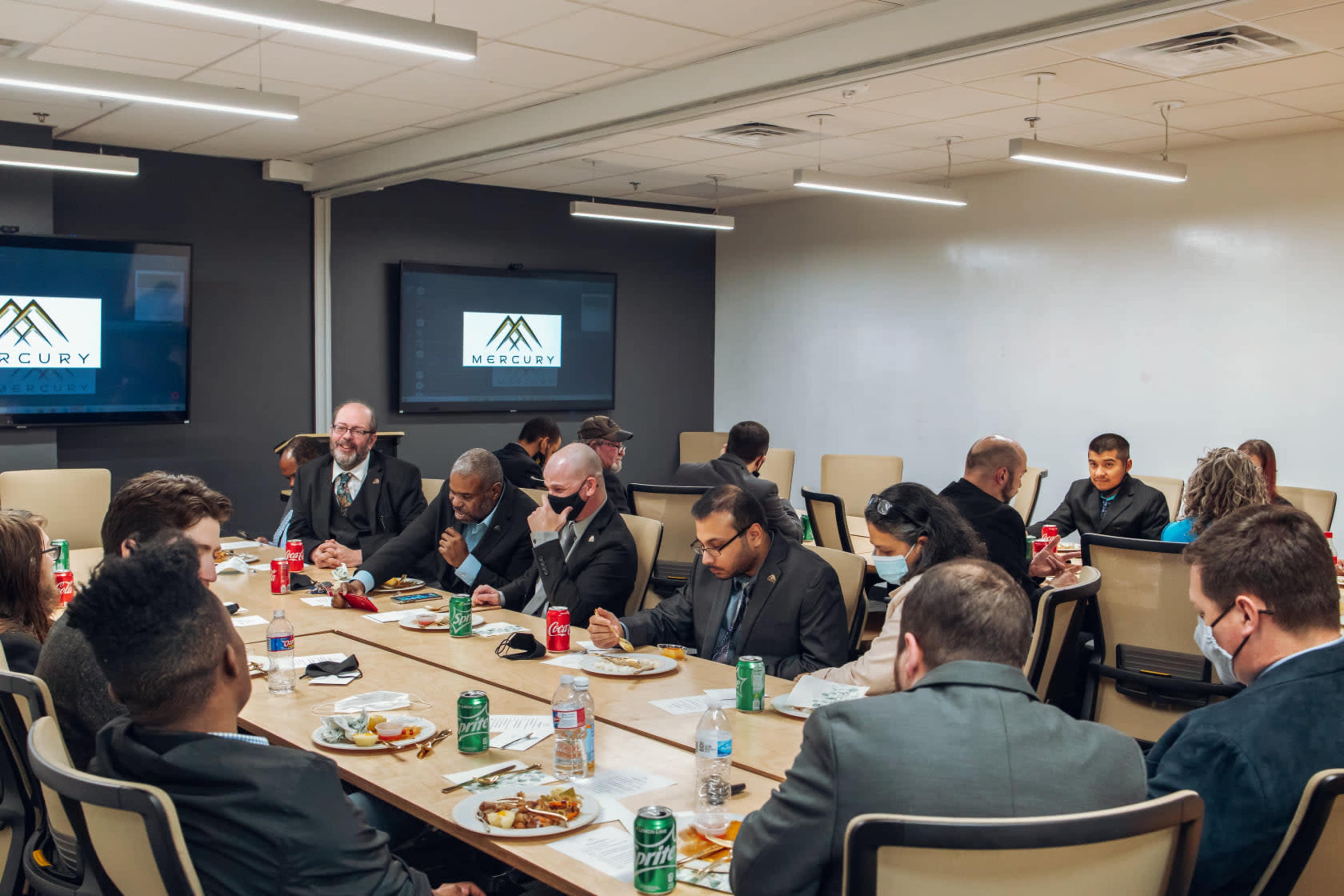 A group of people in business attire are seated around a long table, eating and engaged in conversation during a meal in a conference room.