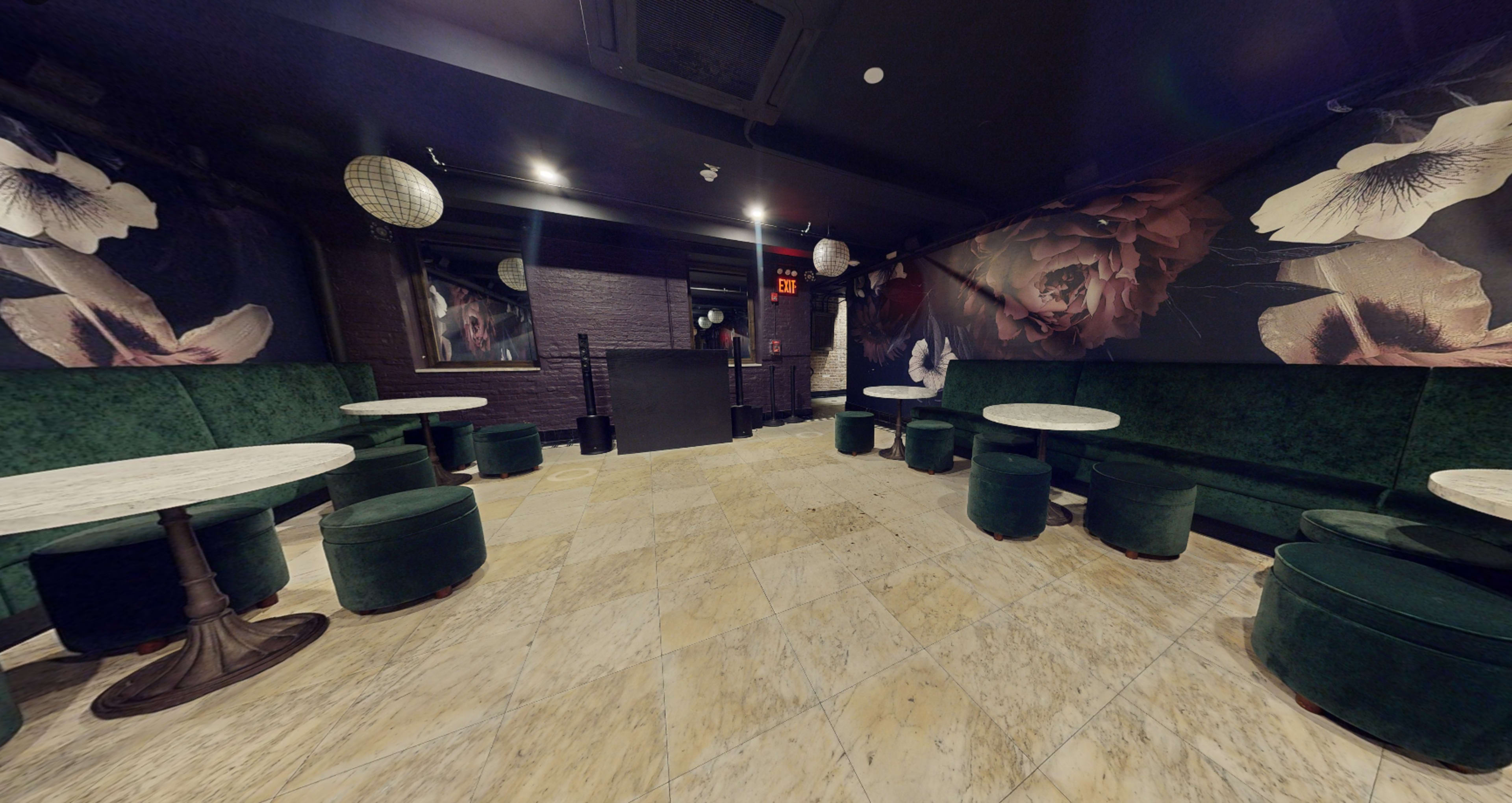 The image depicts a dimly lit lounge area with green velvet seating, marble tables, and floral wall murals.