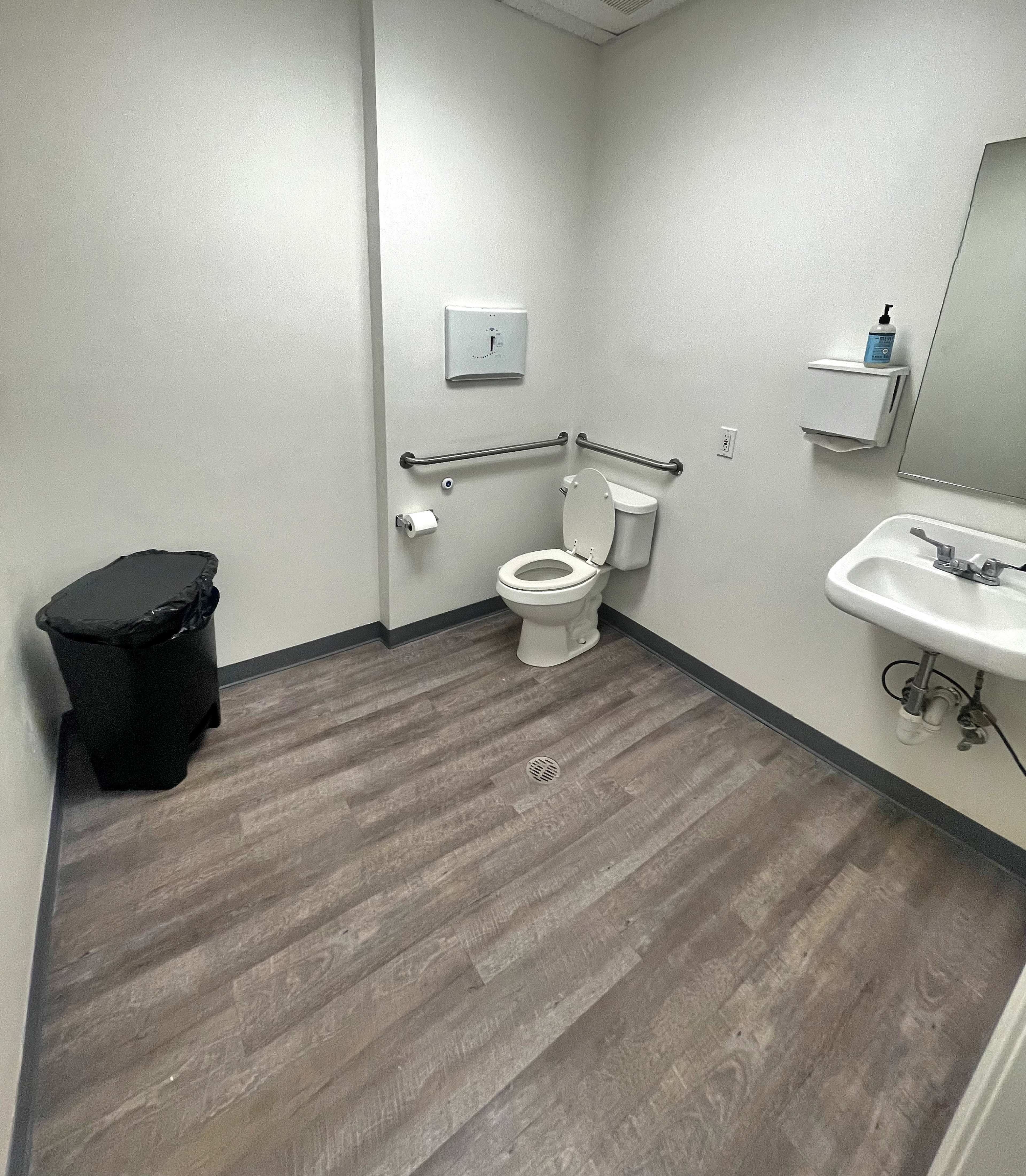 A small restroom features a toilet, sink, trash can, and a mirror, all positioned against plain walls with wood-like flooring.