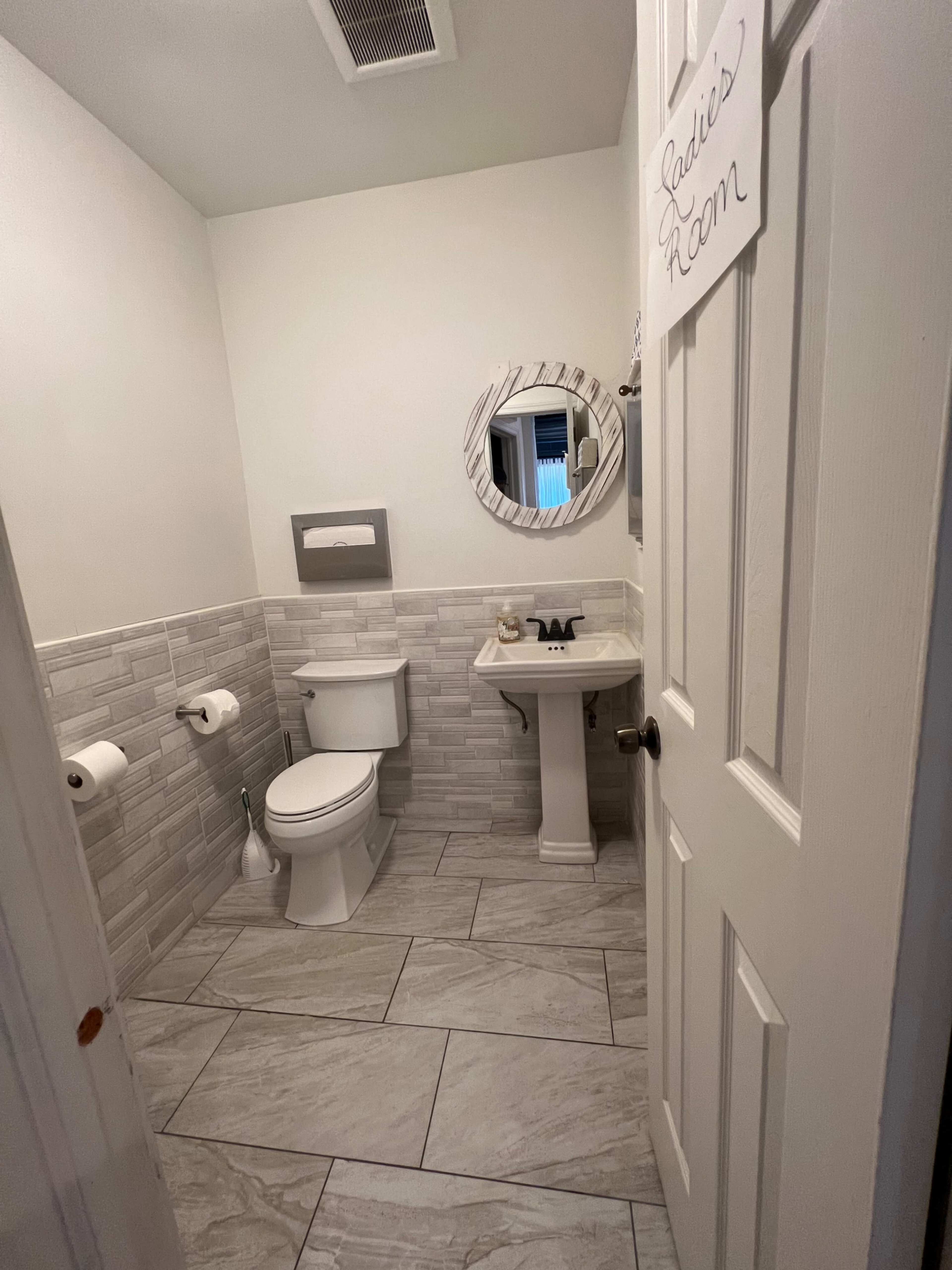 A small bathroom featuring a toilet, sink, and tiled walls with a sign on the door labeling it as "Ladies' Room."