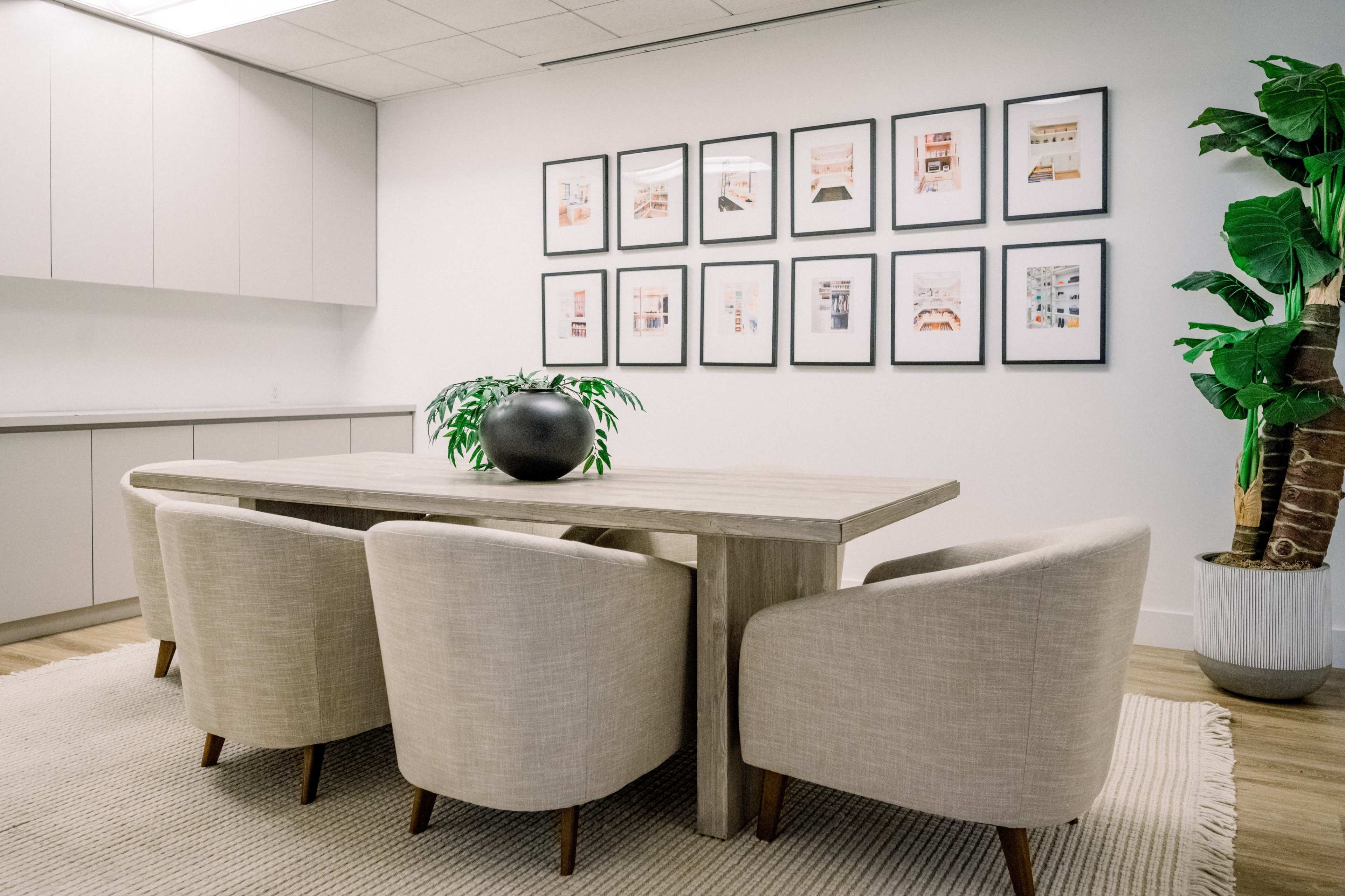 A modern dining area features a wooden table surrounded by four upholstered chairs, with a wall displaying framed artwork and a large potted plant in the corner.