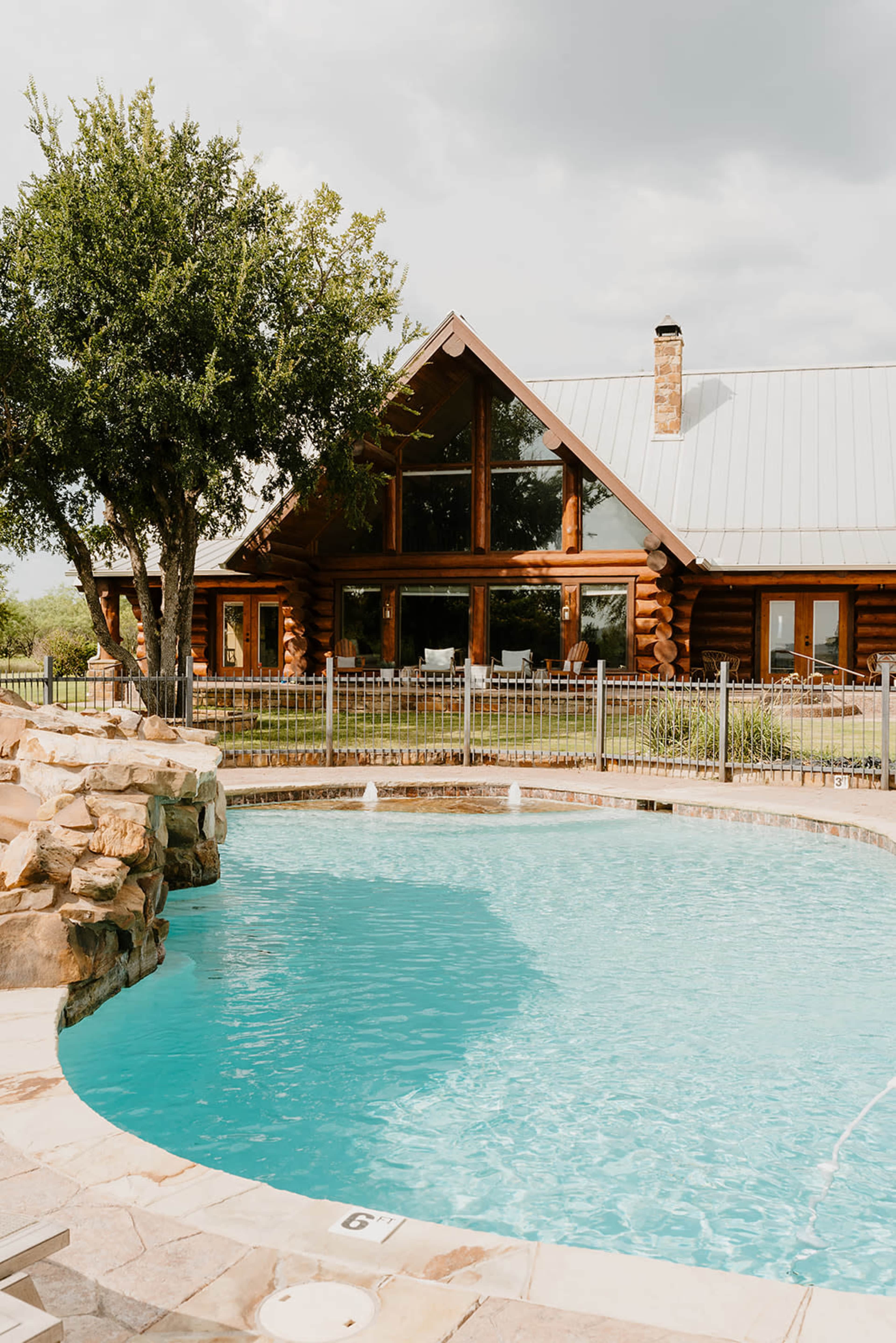 A log cabin with large windows overlooks a blue swimming pool surrounded by a stone patio and lush greenery.