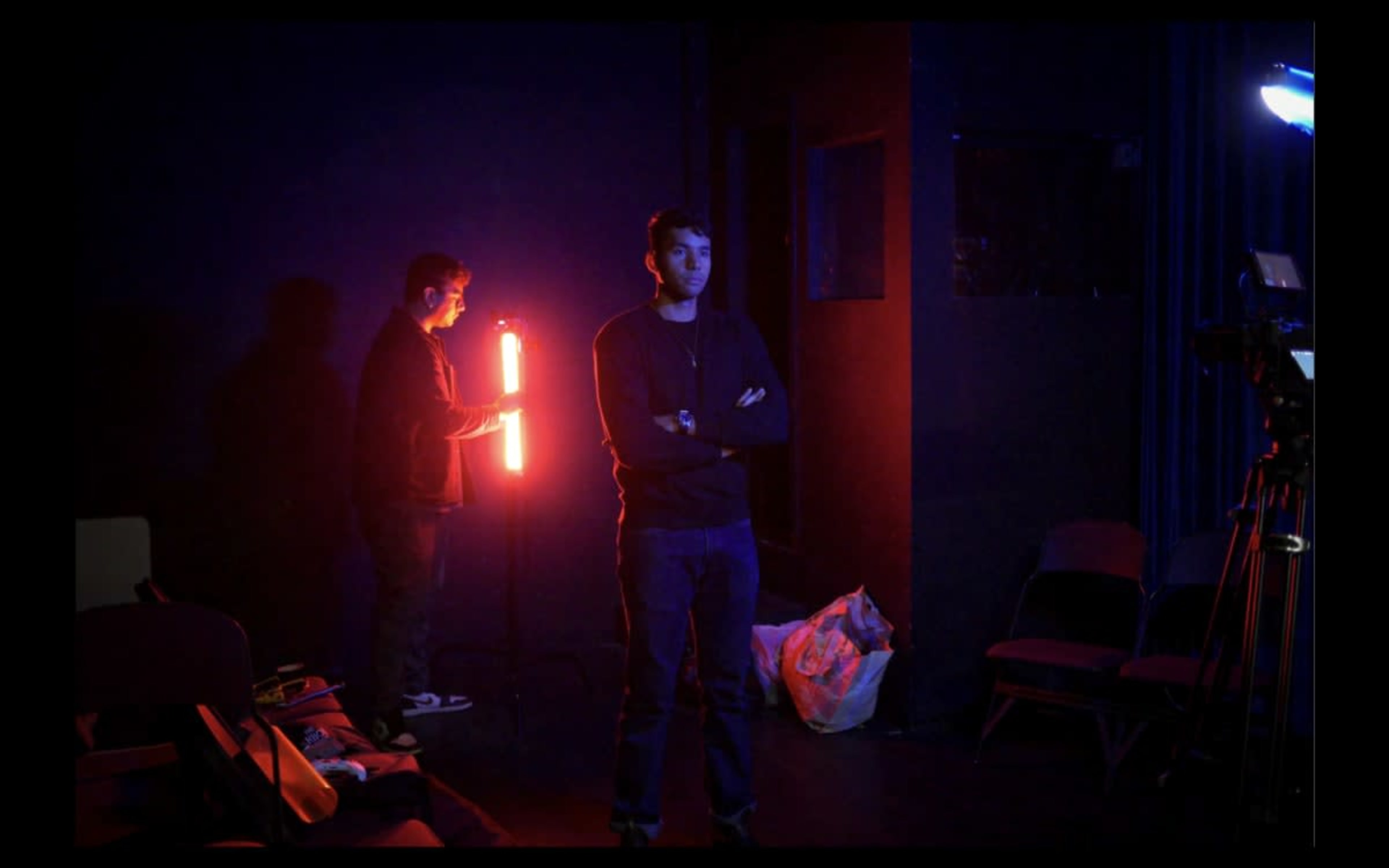 A man stands with his arms crossed in a dimly lit room while another man adjusts a light in the background.