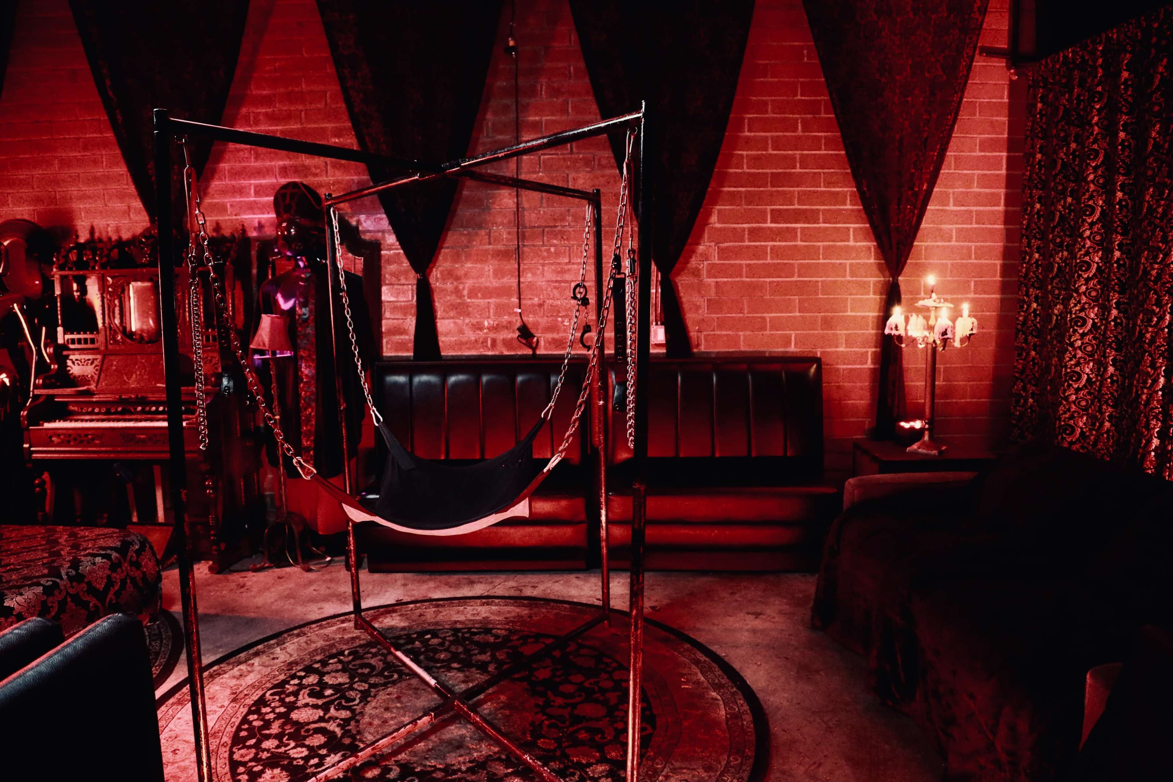 A dimly lit room featuring a metal suspension frame, black couches, and rich red curtains, set against a backdrop of brick walls.