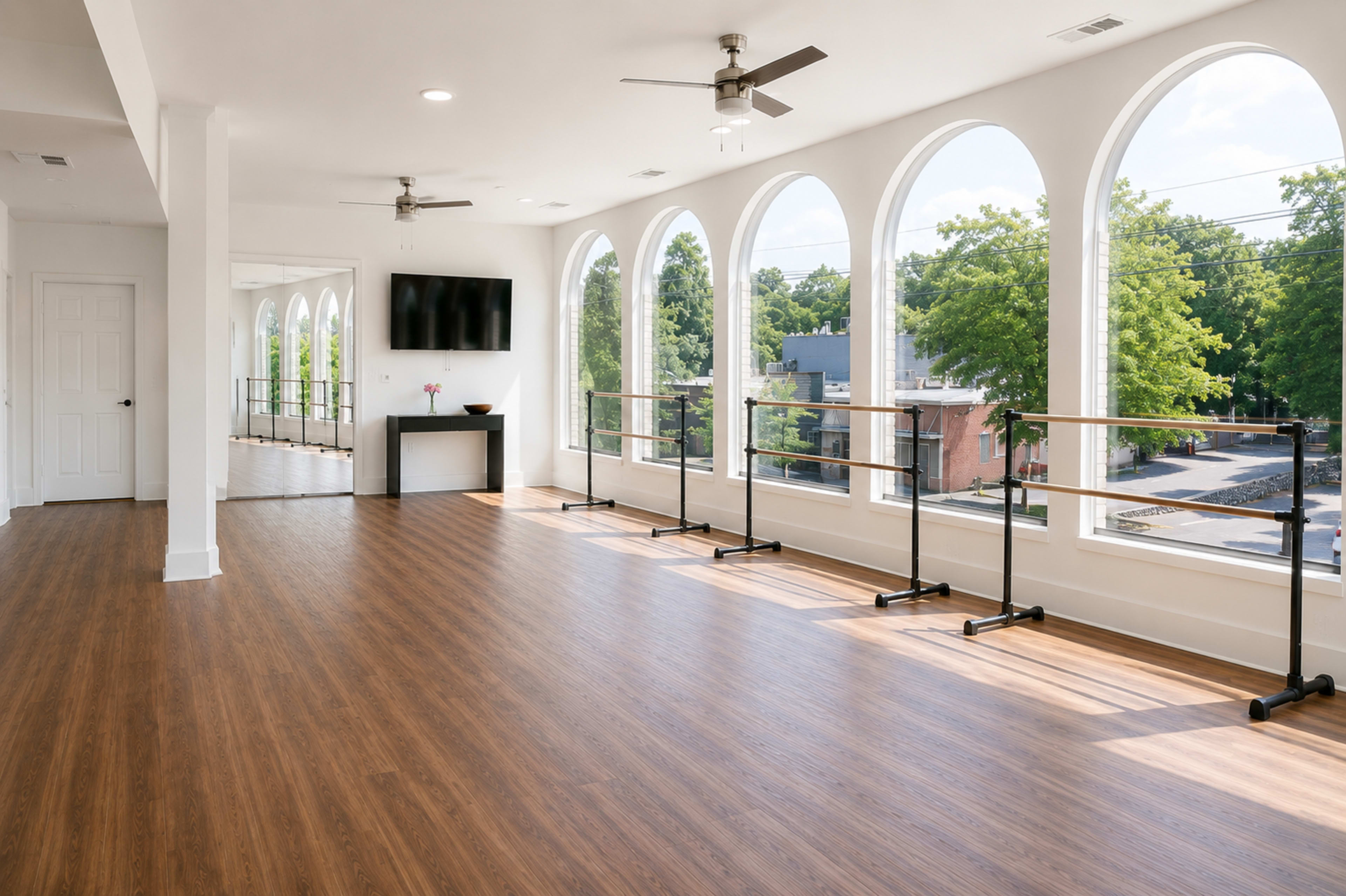 The studio features a spacious interior with wood flooring, large arched windows, ballet barres along the walls, and a television mounted on the wall.