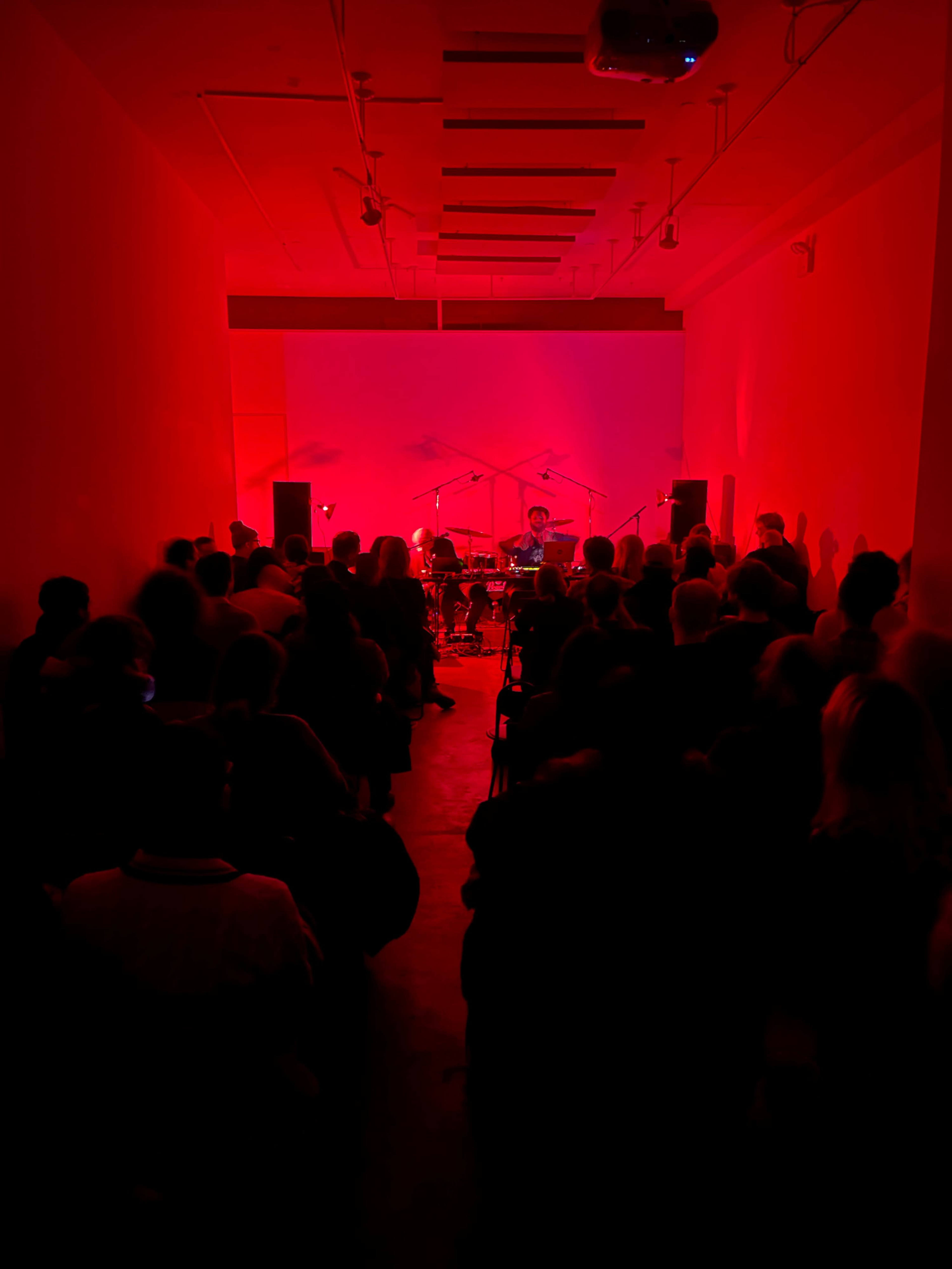 A crowded room with red lighting features a musical performance at the front, highlighting musicians surrounded by microphones and instruments.