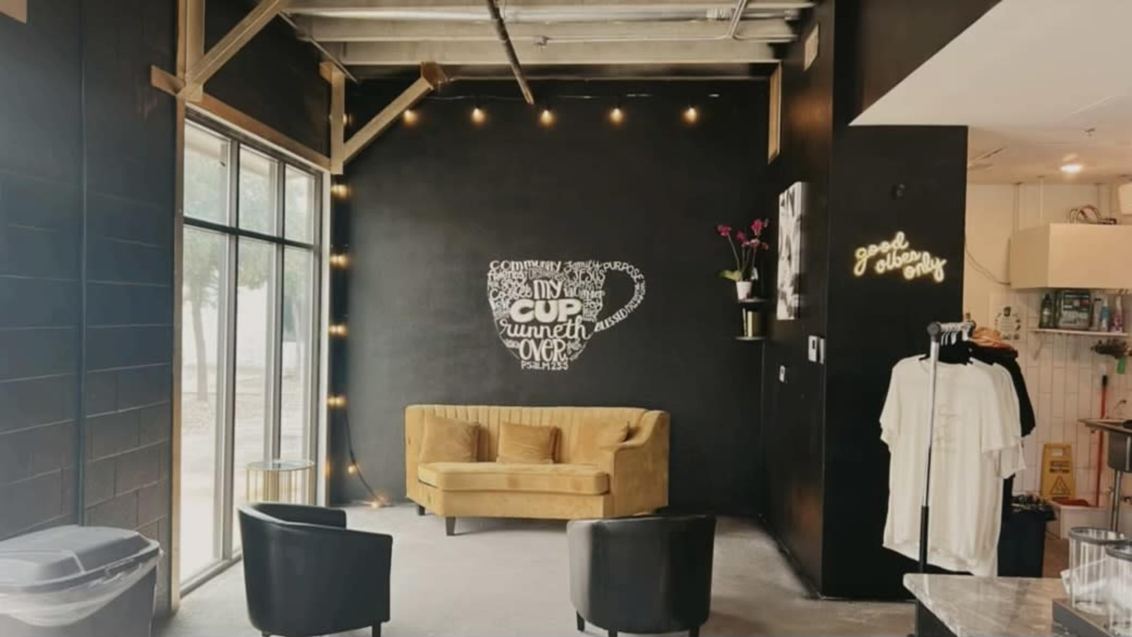 A coffee shop interior with a yellow sofa, a black wall featuring a cup design made of words, and a clothing display on the right.