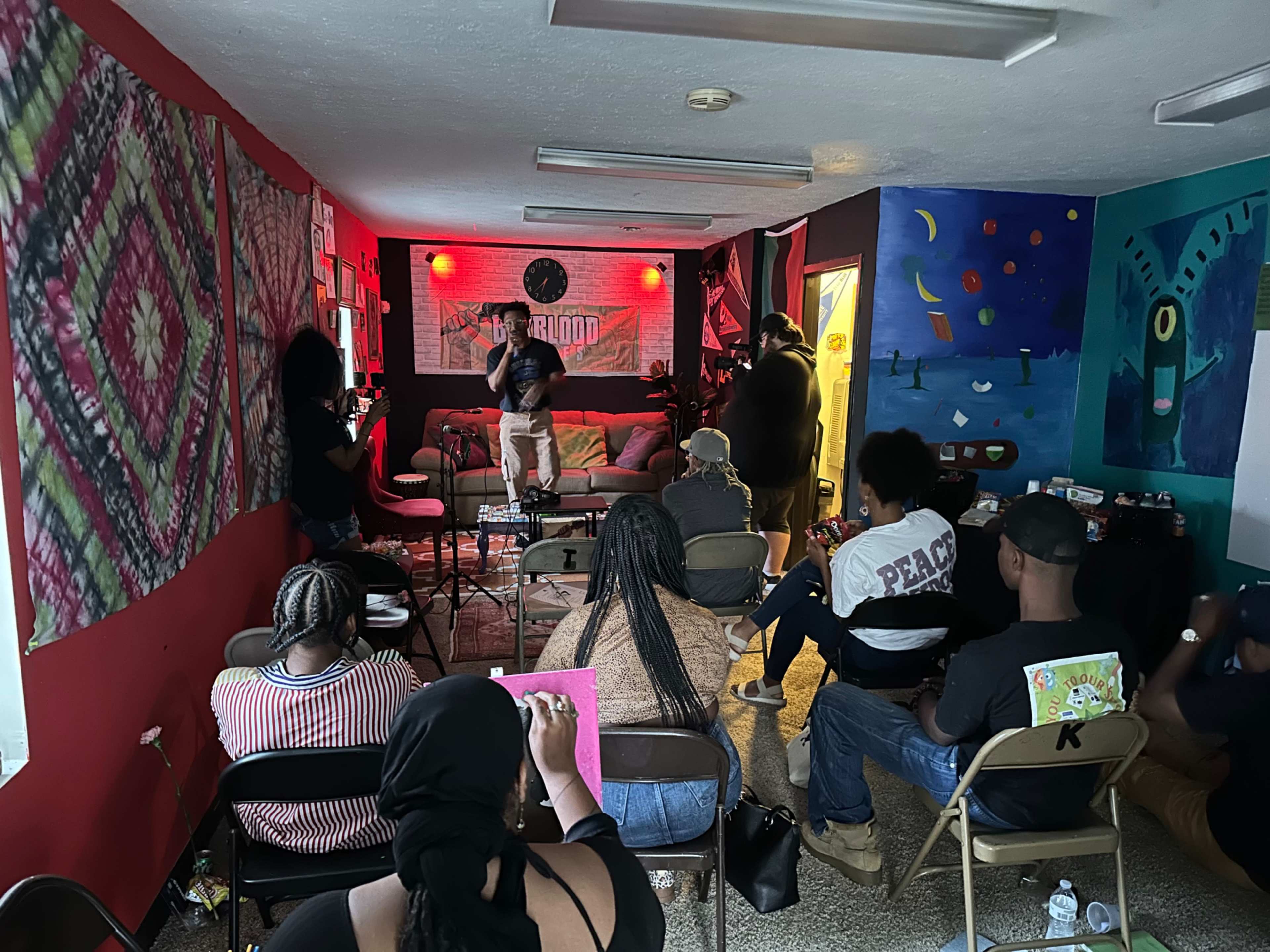 A group of people sits in chairs facing a small stage in a colorful room decorated with various artworks.