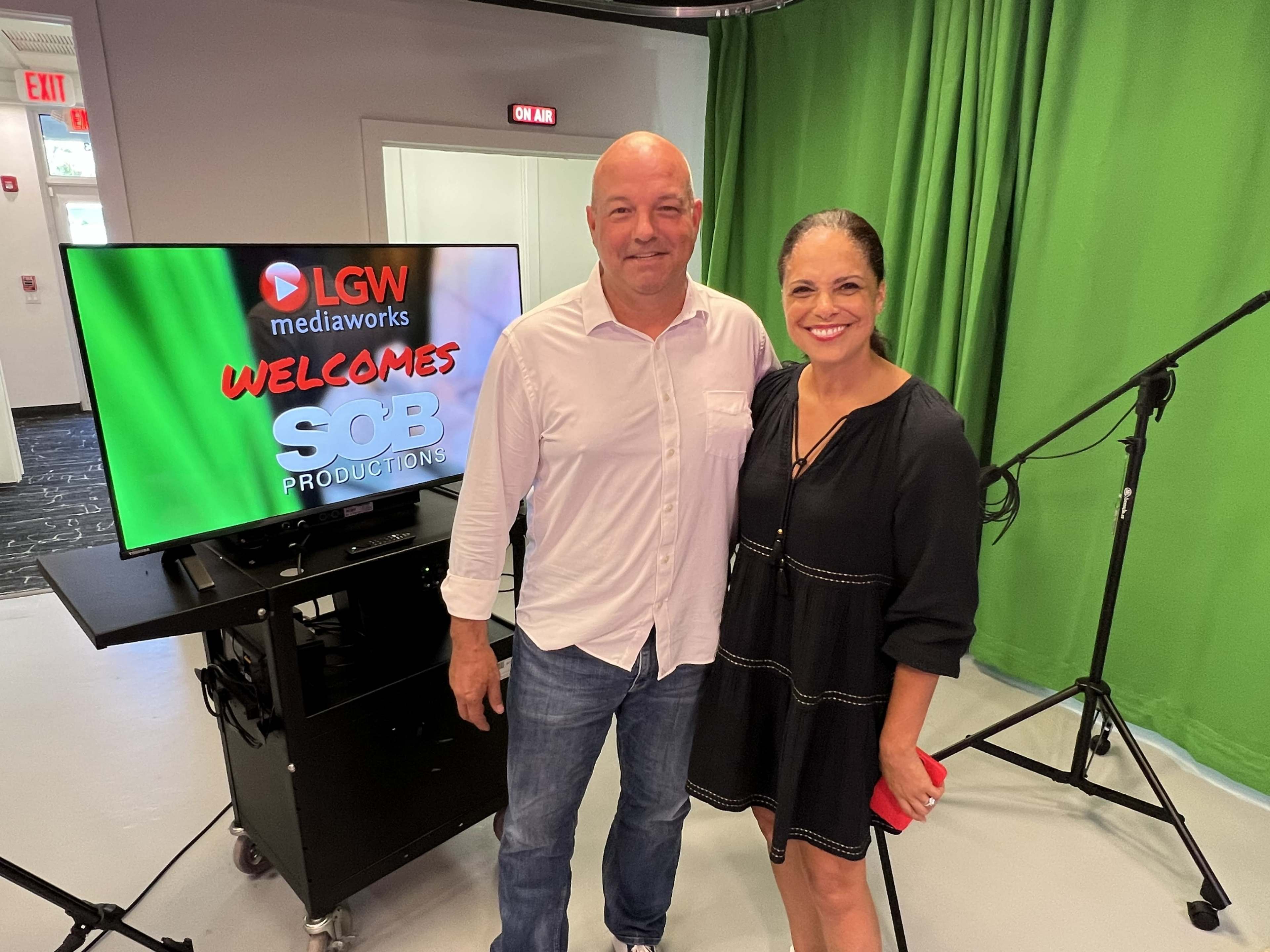 A man and a woman stand together in a studio setting with a green screen backdrop and a monitor displaying "LGW mediaworks WELCOMES SOB PRODUCTIONS."