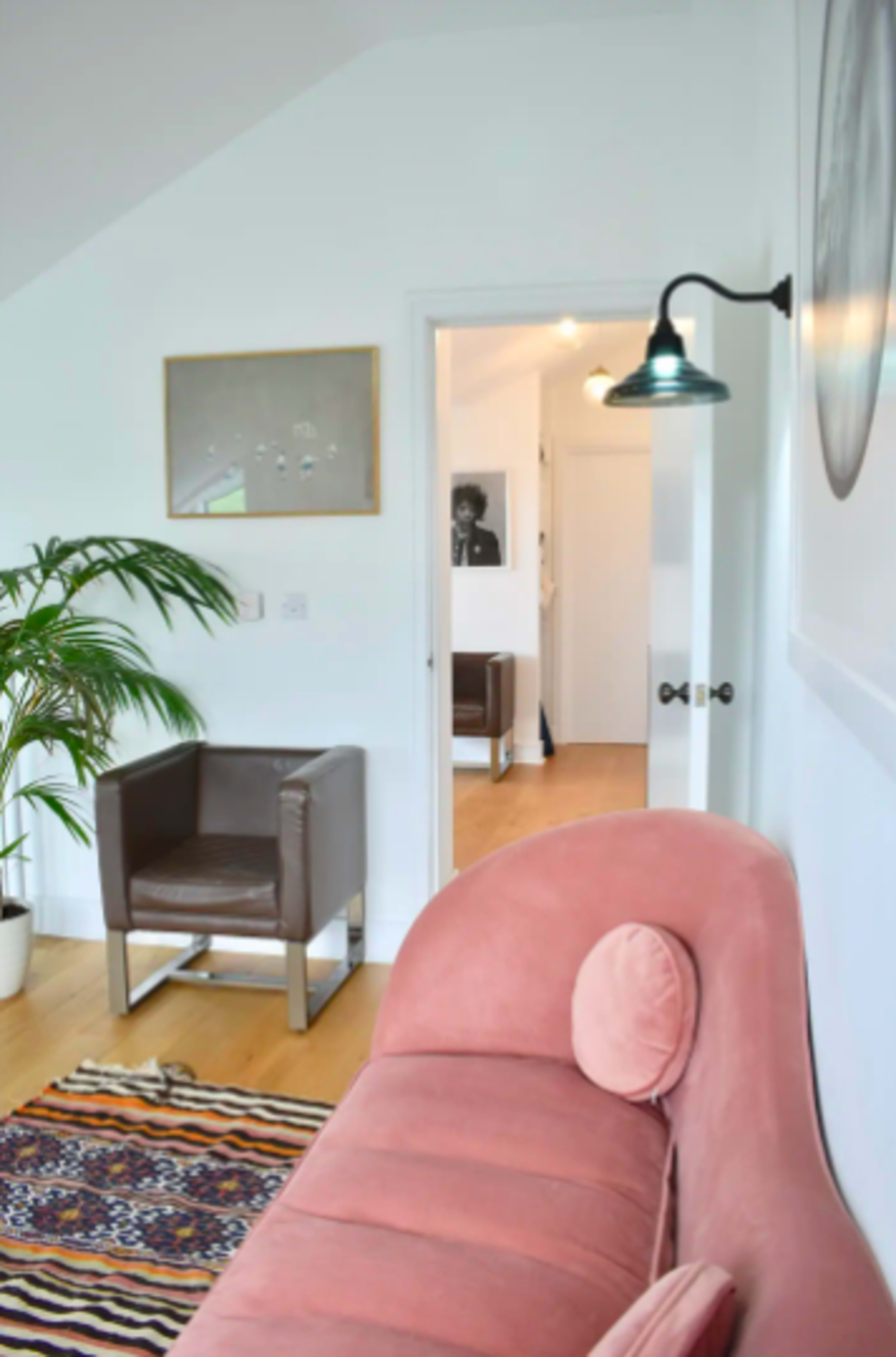 The image shows a modern living room with a pink sofa, a brown armchair, a plant, and a doorway leading to another room.