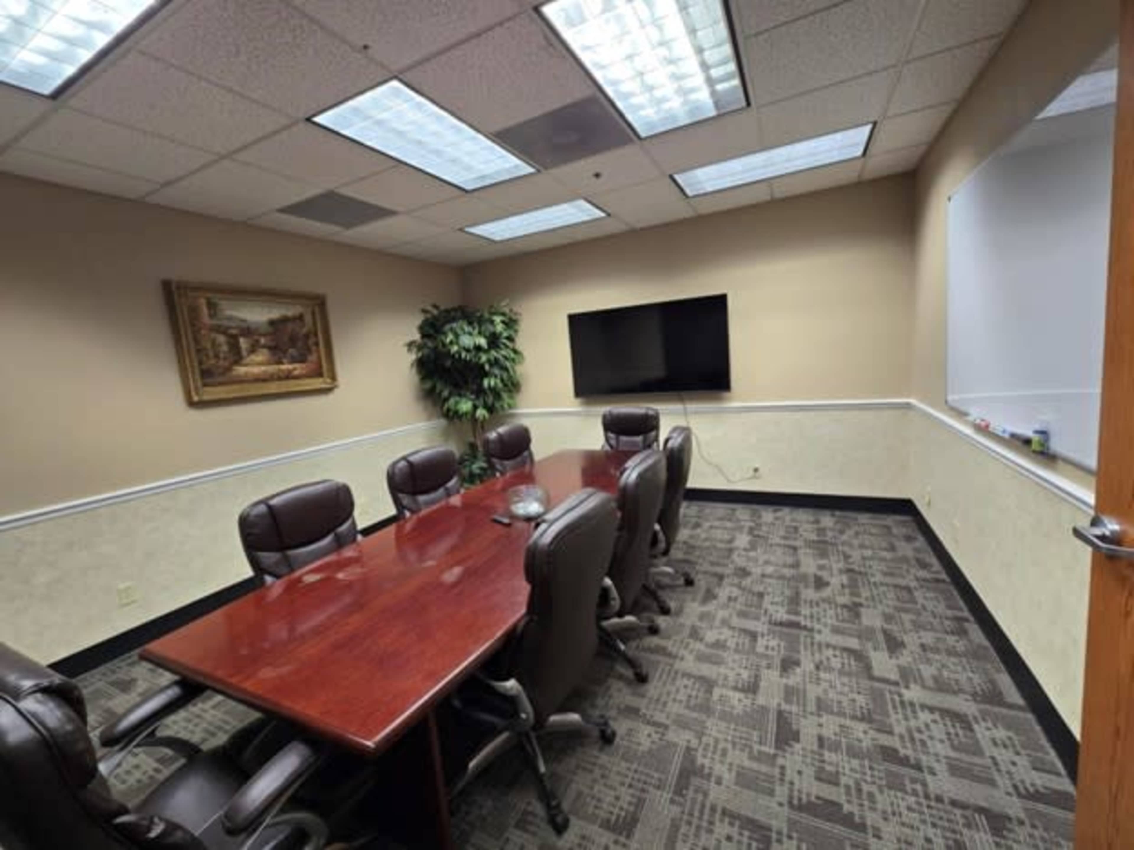 The image shows a conference room with a large wooden table surrounded by chairs, a television mounted on the wall, and a whiteboard in the corner.