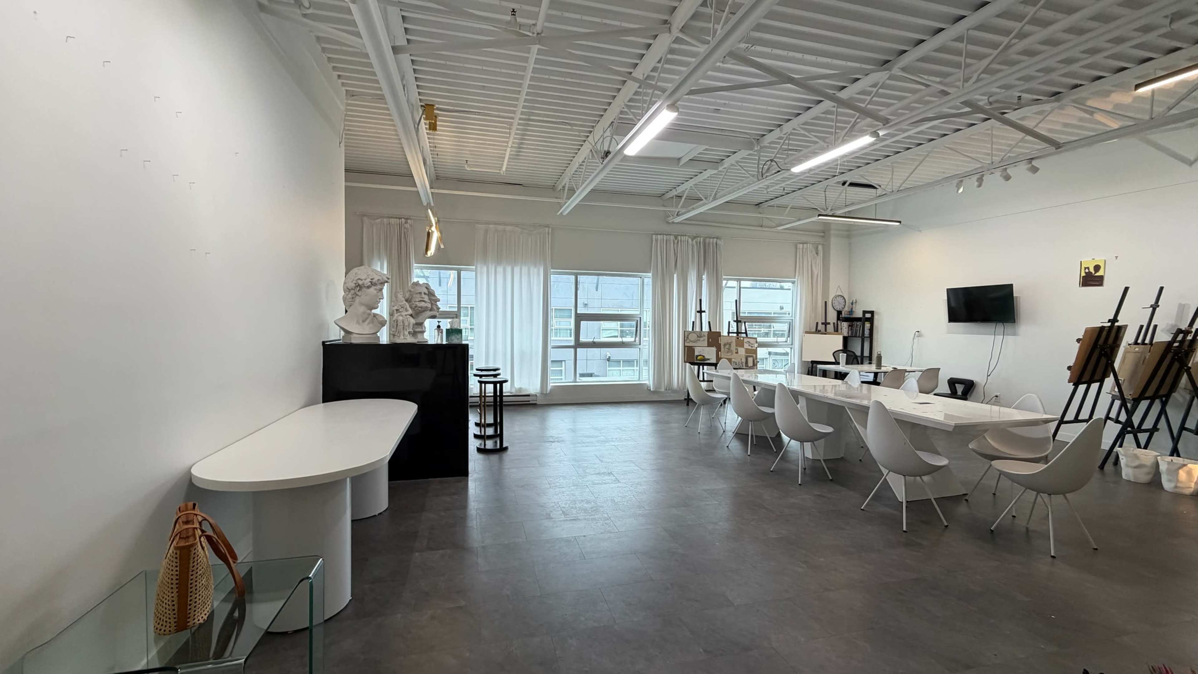 The image shows a spacious, bright art studio featuring white walls, a long dining table, chairs, a few sculptures, and large windows letting in natural light.