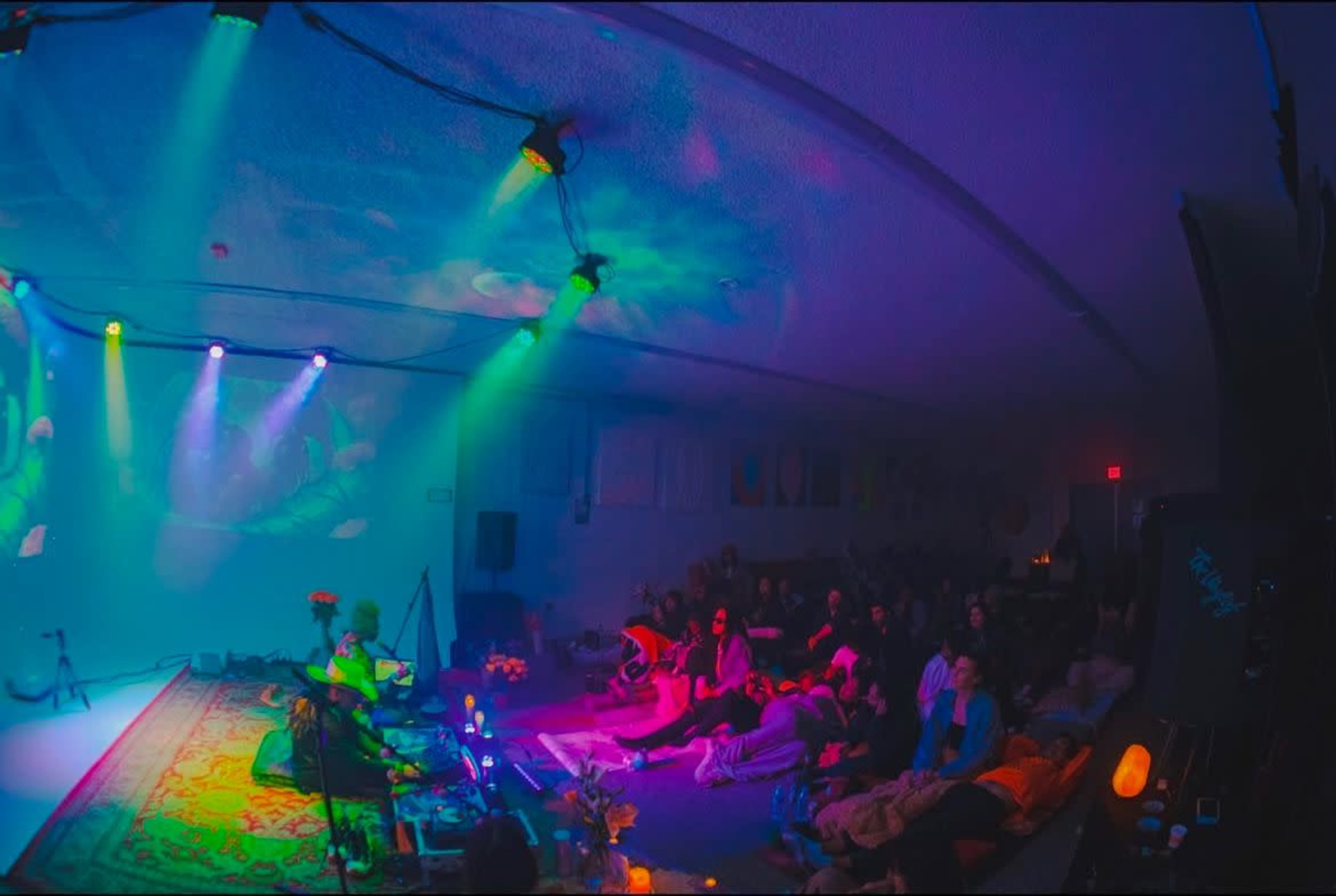 A diverse audience sits on the floor of a dimly lit venue, watching a performer who is playing an instrument under colorful stage lights.