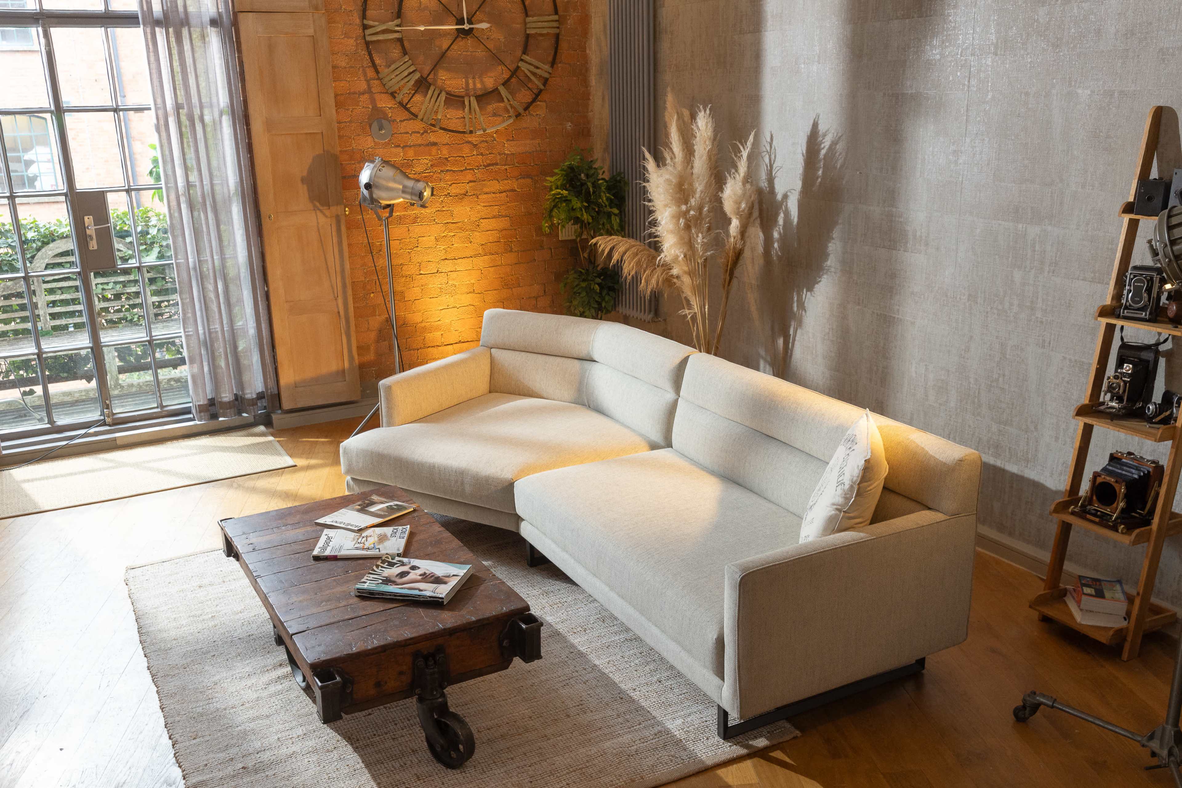 A modern living room features a light-colored sectional sofa, a rustic coffee table, and large windows with sheer curtains, allowing natural light to illuminate the space.